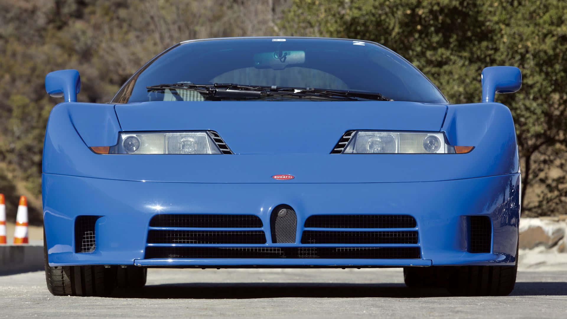 Bugatti Eb110 桌布 壁紙 — 一輛充滿活力的藍色Bugatti EB110超級跑車在陽光明媚的道路上向前行駛，背景有樹木和橙色錐體。