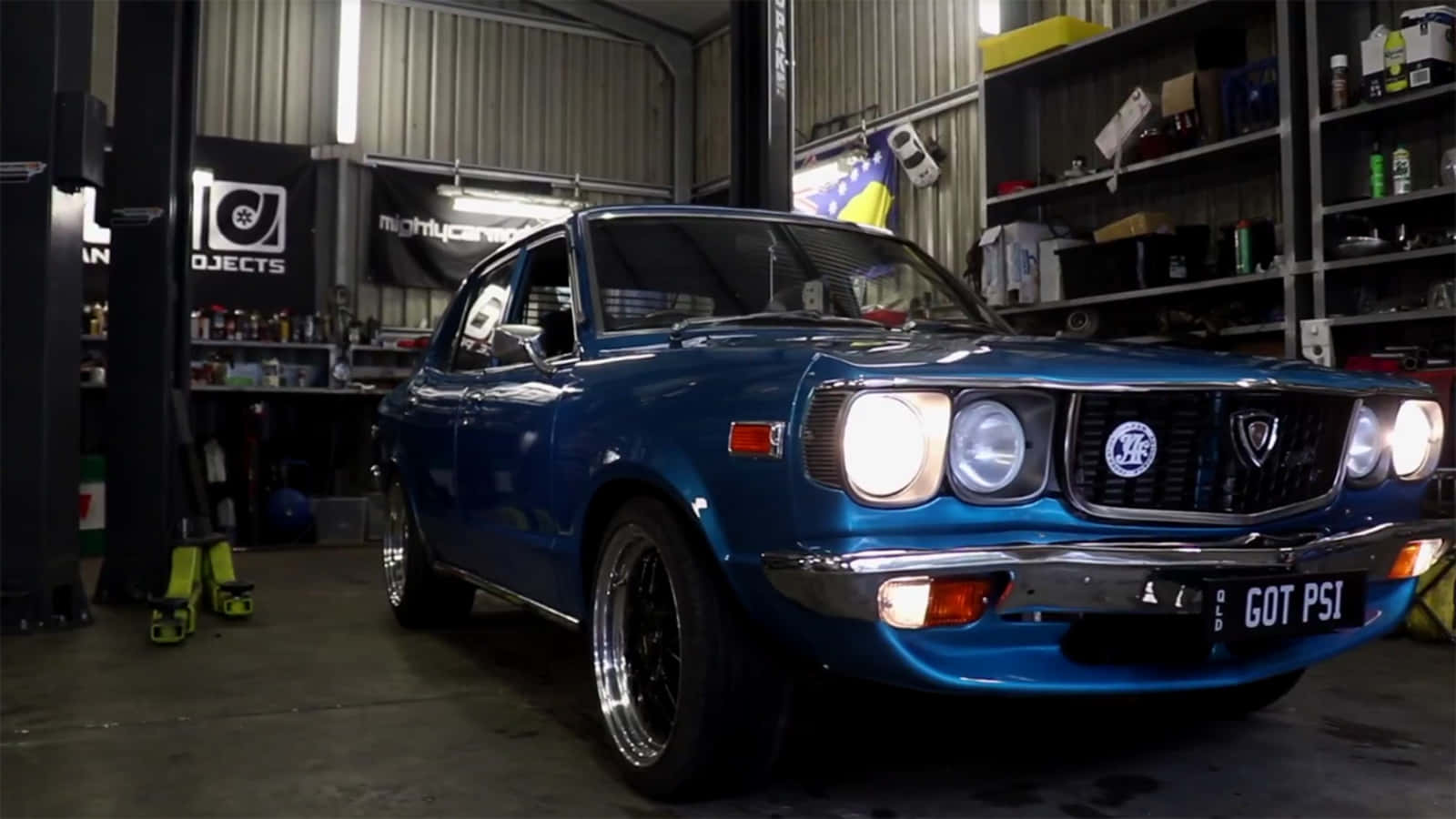 Mazda Rx-3 桌布 壁紙 — 一輛充滿活力的藍色經典汽車，車頭燈閃閃發光，自豪地停在一個雜亂的車庫裡，車庫裡裝滿了工具和汽車紀念品。