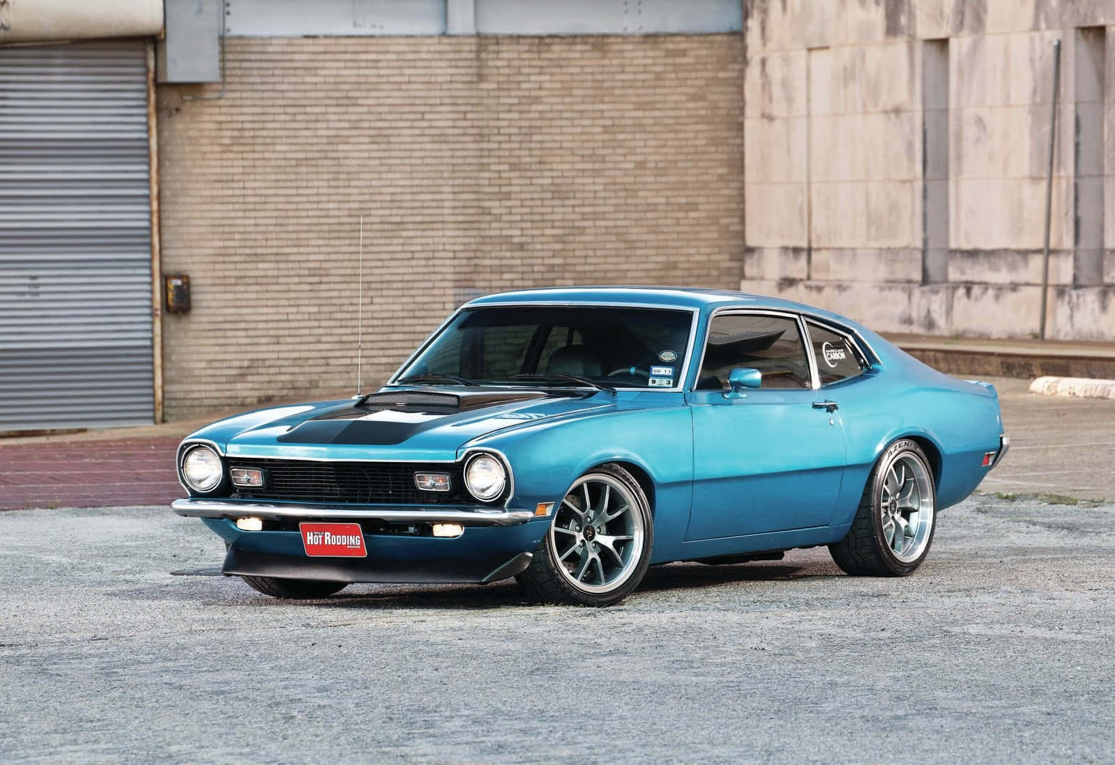 Ford Maverick 桌布 壁紙 — 一輛充滿活力的藍色經典肌肉車，帶有黑色引擎蓋條紋，停在磚砌建築前的礫石上。