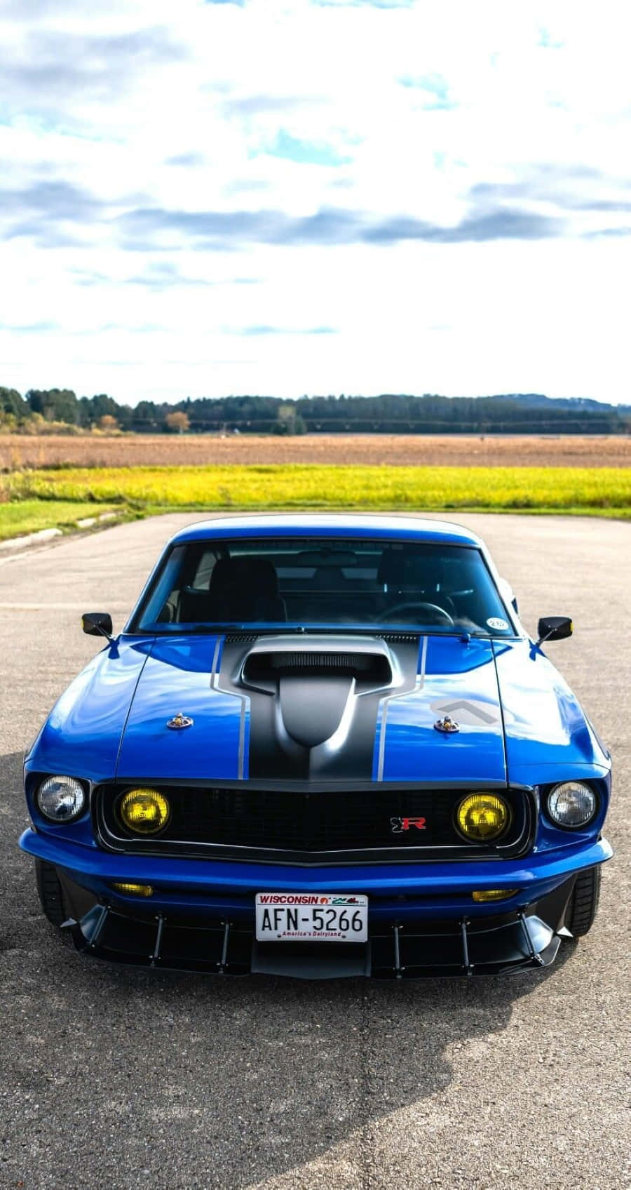 Ford Mustang Mach 1 桌布 壁紙 — 一輛充滿活力的藍色經典肌肉車，帶有黑色賽車條紋，自豪地停在鋪砌的道路上，背景是金色的田野和多雲的天空。