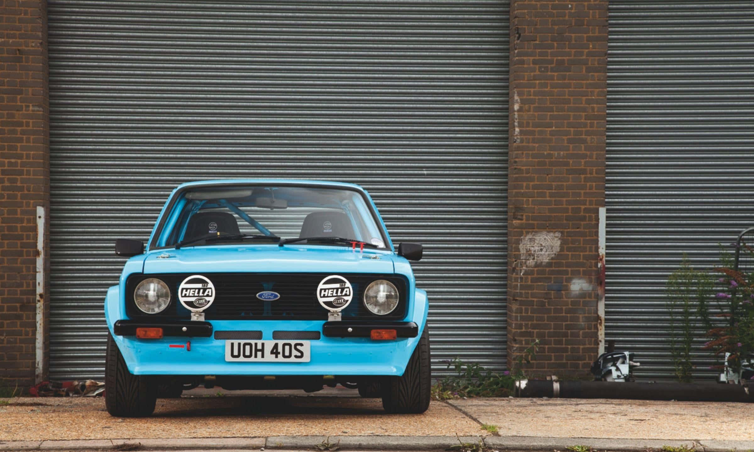 Ford Escort 桌布 壁紙 — 一輛鮮藍色經典拉力賽車，配備Hella車燈，停泊在波紋金屬車庫門前。
