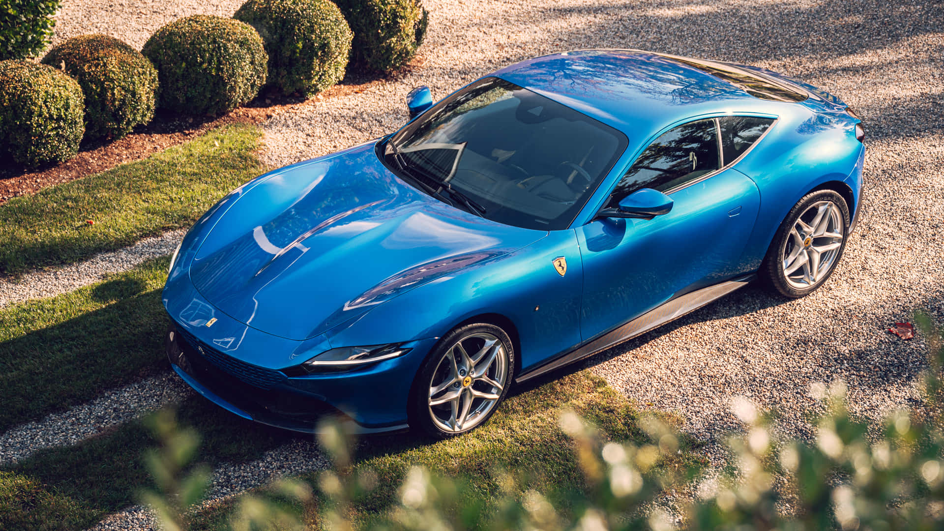 Ferrari Roma 桌布 壁紙 — 一輛充滿活力的藍色法拉利跑車在陽光下閃閃發光，停在碎石車道上，旁邊是修剪整齊的綠色樹籬。