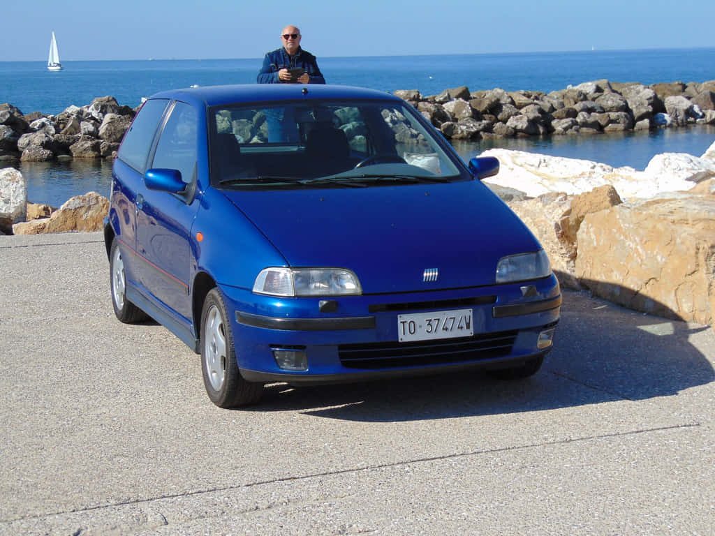 Fiat Punto 桌布 壁紙 — 一輛鮮藍色的Fiat Punto停泊在陽光普照的海岸公路上，一名男子站在車頂，地平線上可見一艘帆船。