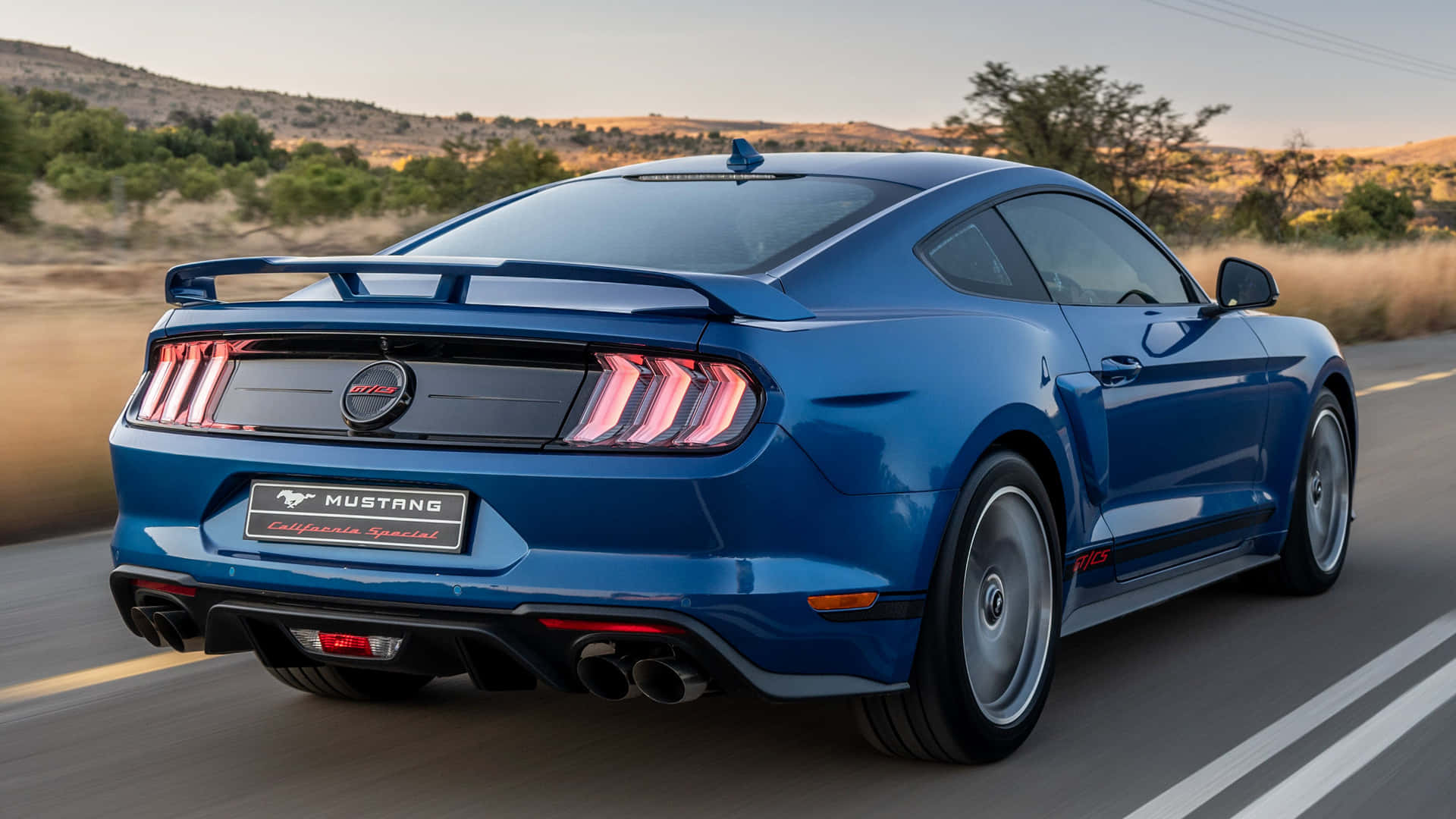 Ford Mustang California Special 桌布 壁紙 — 一輛充滿活力的藍色福特野馬加州特別版在日落時分沿著沙漠公路行駛，尾燈閃爍發光。