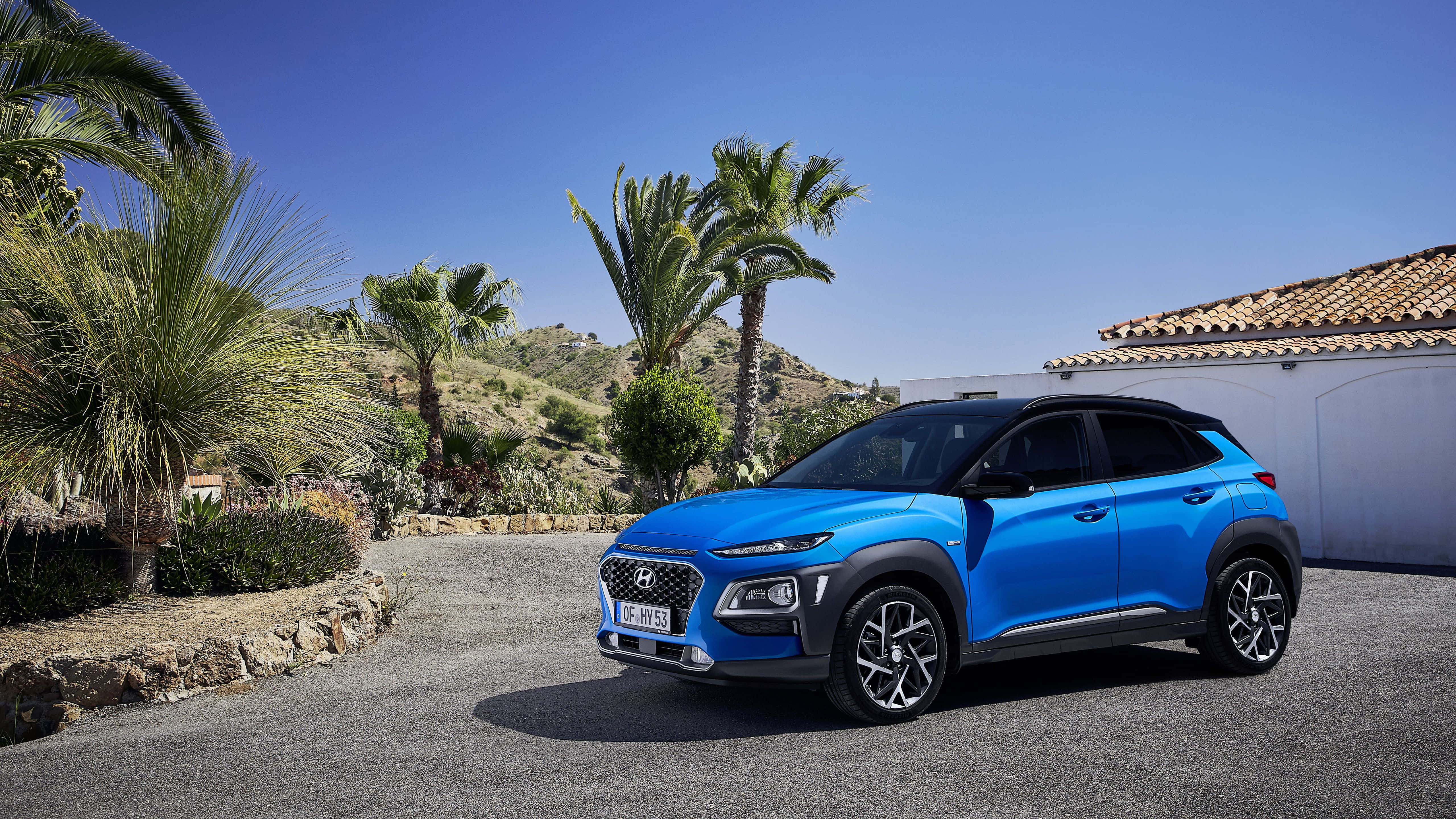 Hyundai Kona 桌布 壁紙 — 一輛充滿活力的藍色現代Kona SUV停放在陽光普照的車道上，旁邊有棕櫚樹和白色建築，背景是清澈的藍天。