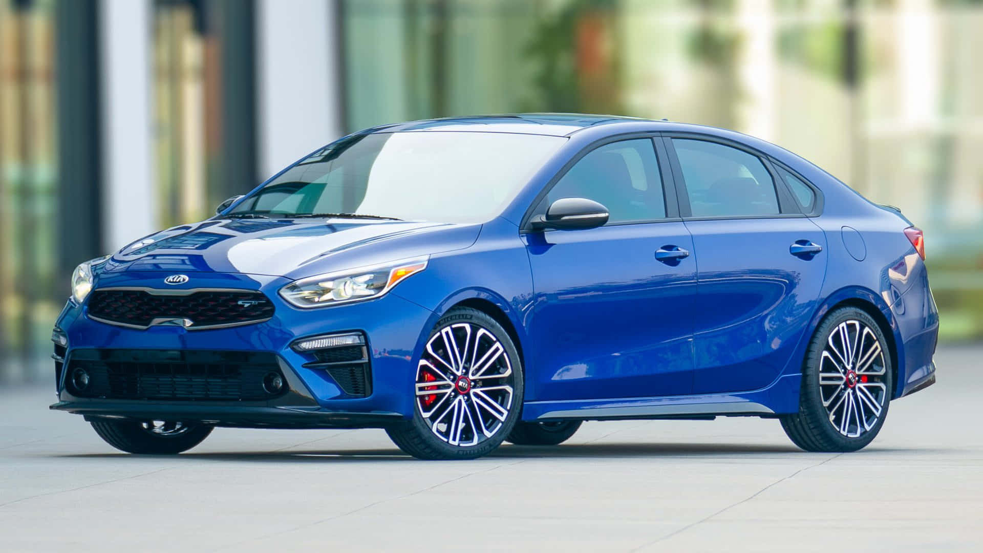 Kia Forte 桌布 壁紙 — 一輛充滿活力的藍色Kia轎車，配備紅色制動卡鉗和時尚合金輪圈，停放在現代城市人行道上。