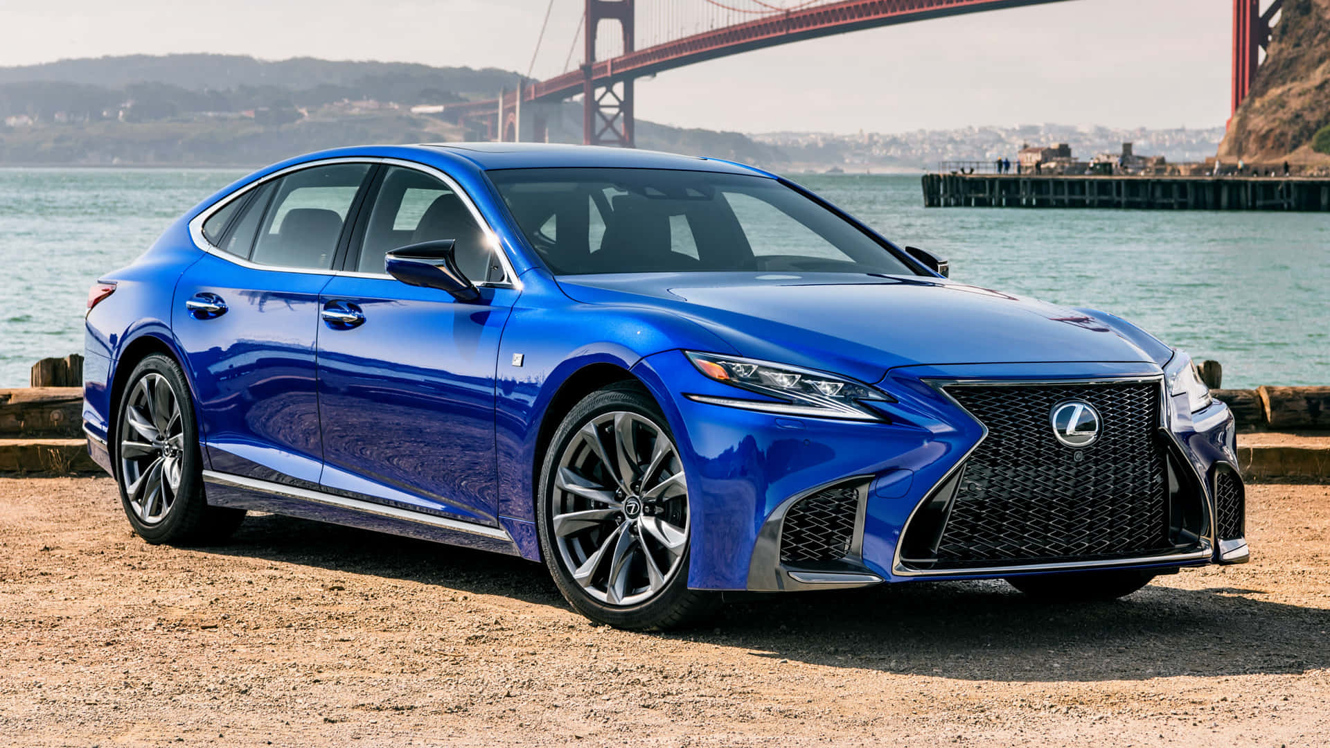 Lexus Ls 500 桌布 壁紙 — 一輛充滿活力的藍色Lexus房車在陽光下閃閃發光，停在碎石岸邊，背後是雄偉的金門大橋。