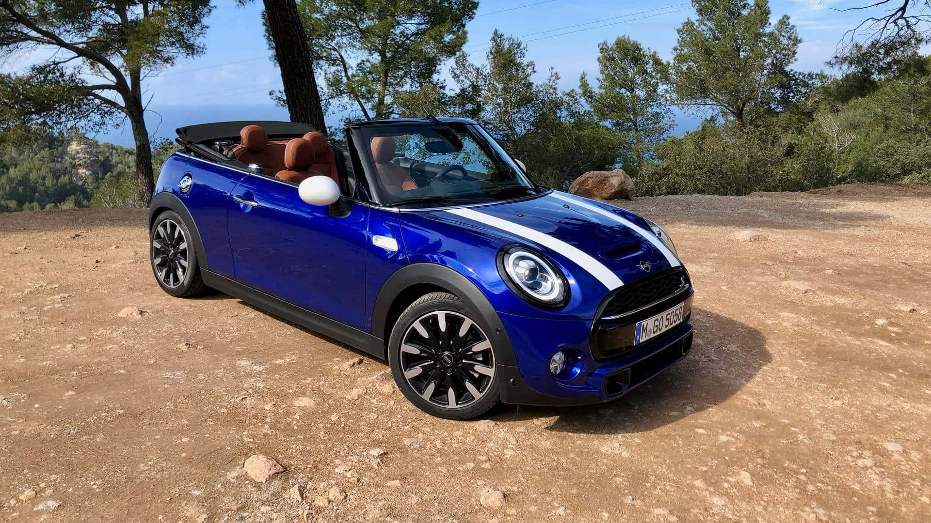 Mini Cooper S Convertible 桌布 壁紙 — 一輛充滿活力的藍色MINI Cooper敞篷車停放在陽光明媚的泥土小徑上，背景有松樹和海洋景觀。