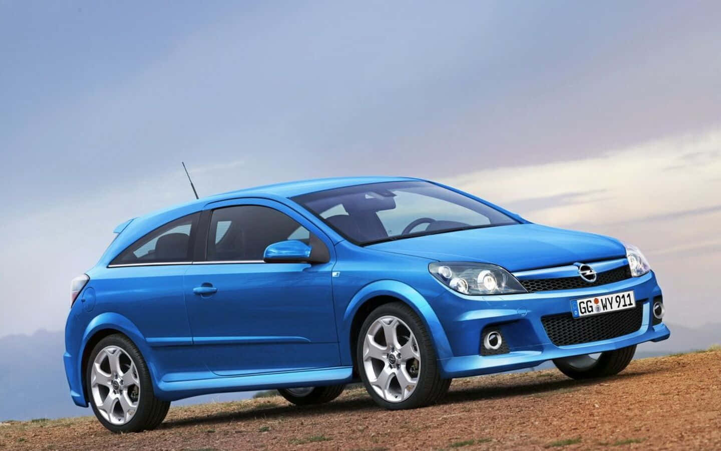 Opel Astra 桌布 壁紙 — 一輛充滿活力的藍色歐寶雅特 GTC 跑車停放在泥土山坡上，天空呈現柔和暮色。