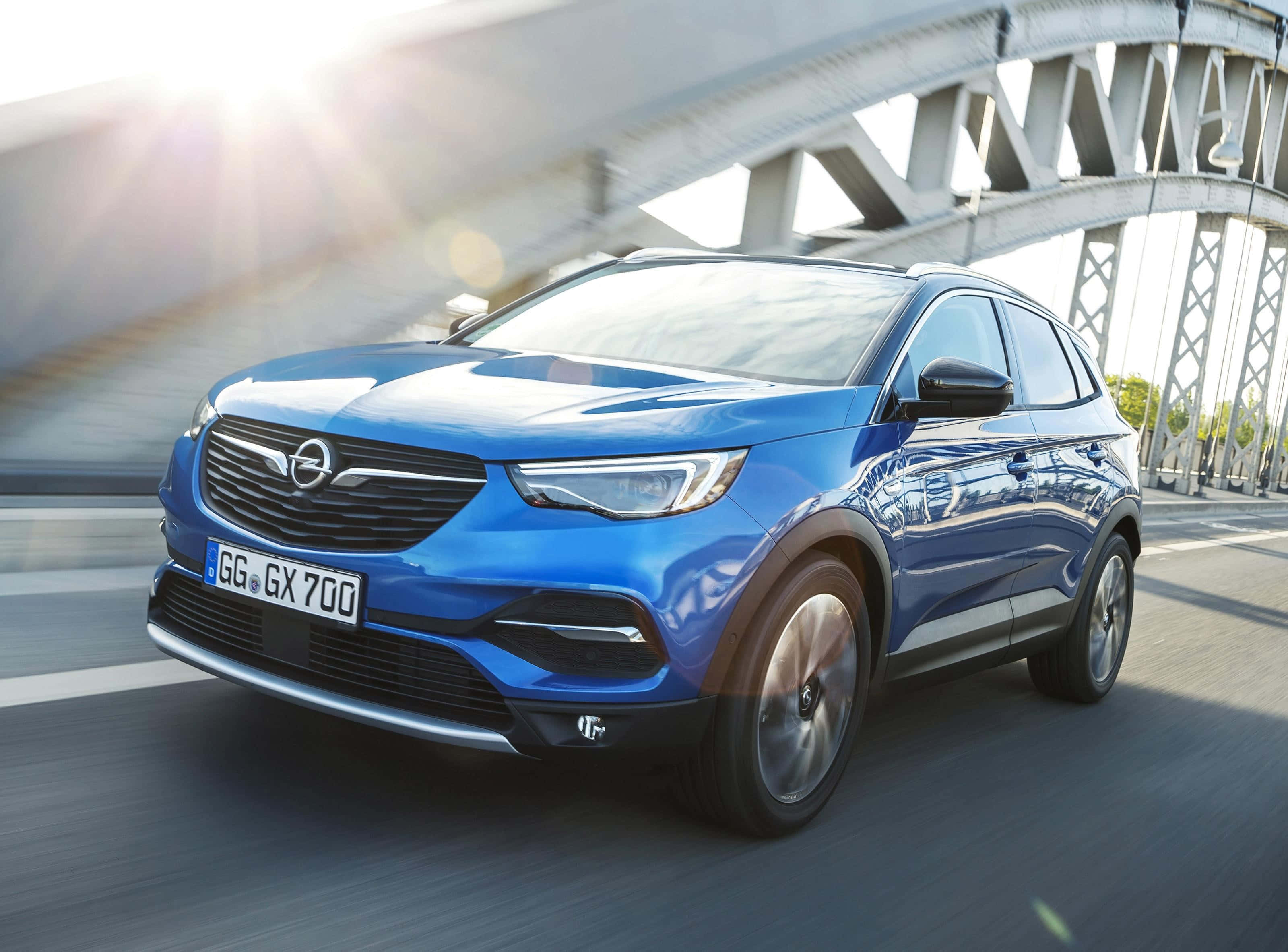 Opel Grandland X 桌布 壁紙 — 一輛充滿活力的藍色歐寶SUV在鋼橋下疾馳，伴隨著太陽耀斑和道路上的動態模糊。