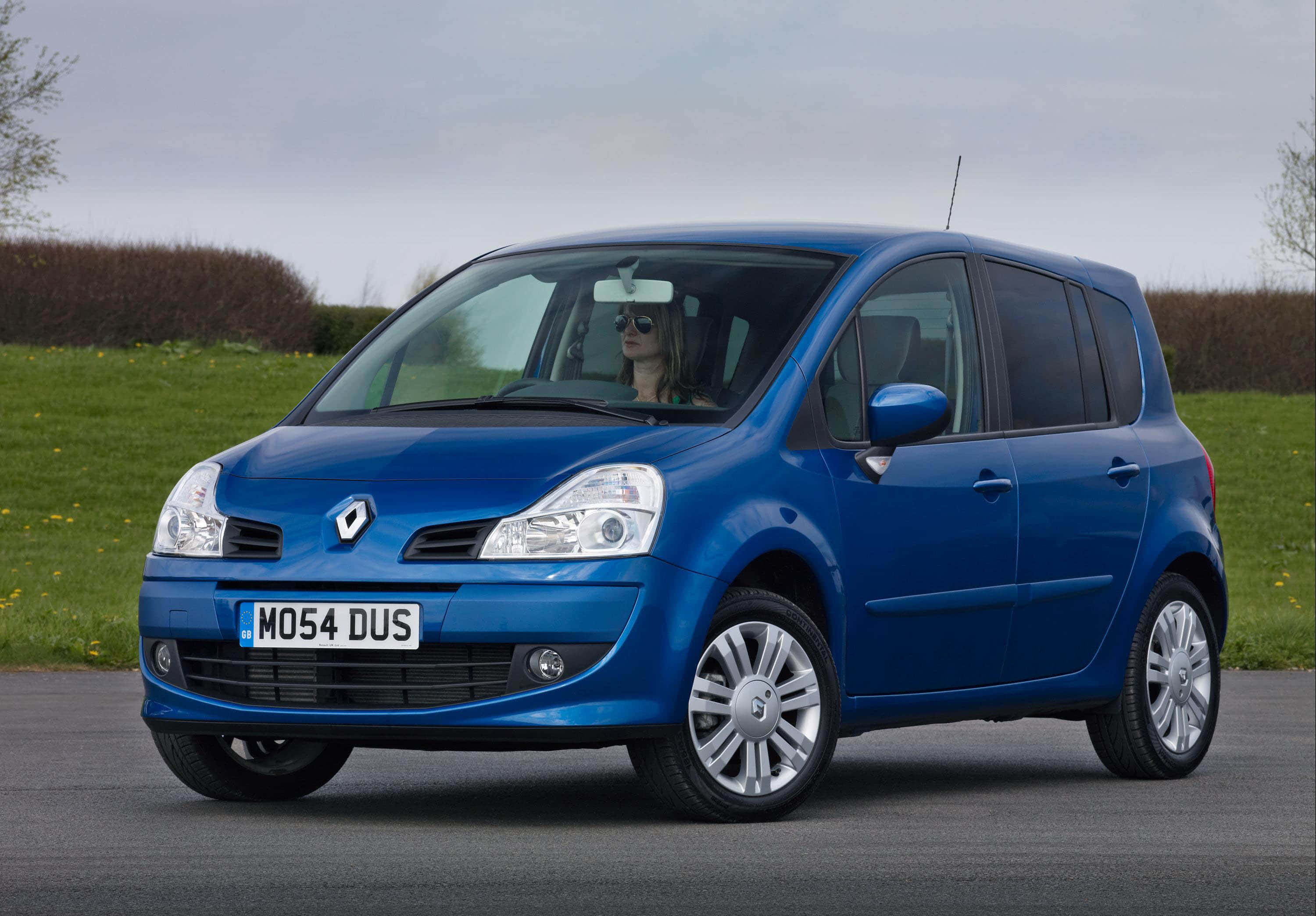 Renault Modus 桌布 壁紙 — 一輛充滿活力的藍色雷諾 Grand Scenic 停在瀝青路上，一名戴著太陽眼鏡的女性坐在方向盤後面，背景是草地田野。