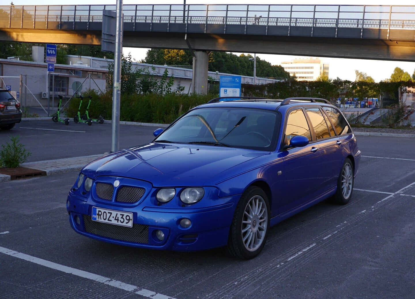 Mg Zt 桌布 壁紙 — 一輛充滿活力的藍色旅行車在黃金時段停在大橋下的停車場，溫暖的陽光反射在其光滑的表面上。