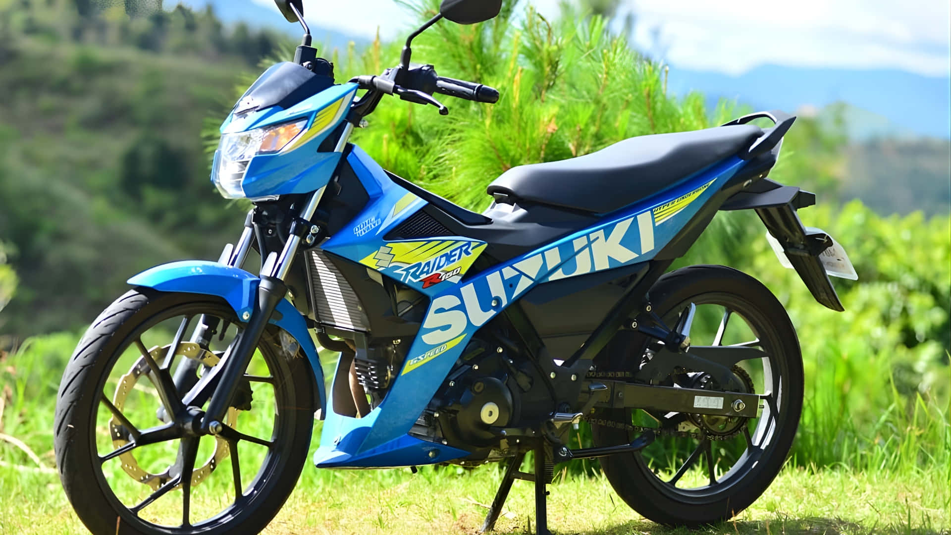  壁紙 — 一輛鮮藍色的Suzuki Raider 150機車停在草地上，背景是翠綠的山丘和遠處的山脈，天空明亮。