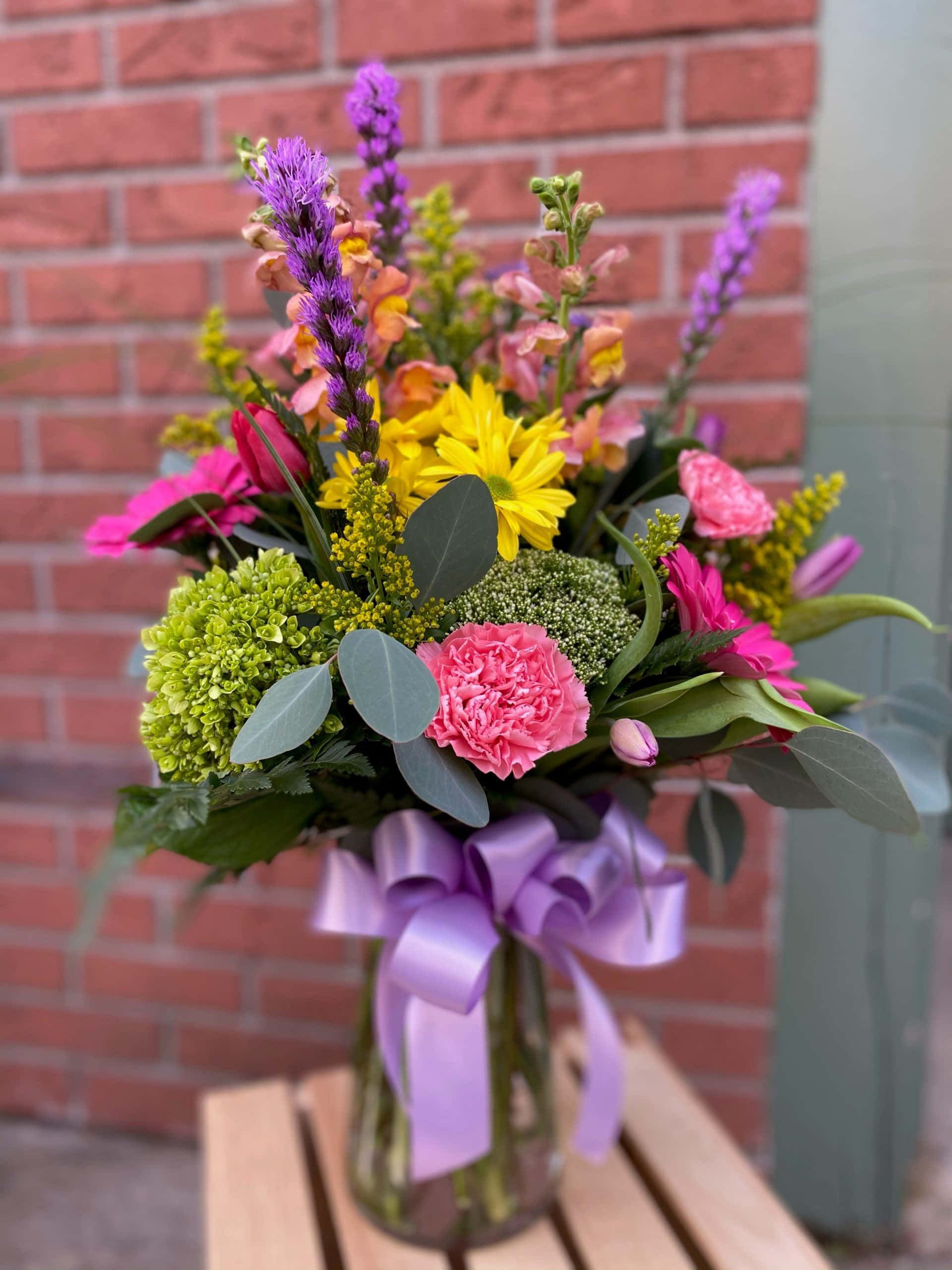 Flower Arrangement 桌布 壁紙 — 一個充滿活力的花束，包含粉紅色康乃馨、黃色雛菊和紫色穗狀花，放在一個玻璃花瓶裡，繫著淡紫色蝴蝶結，置於木桌上，背景是紅磚牆。