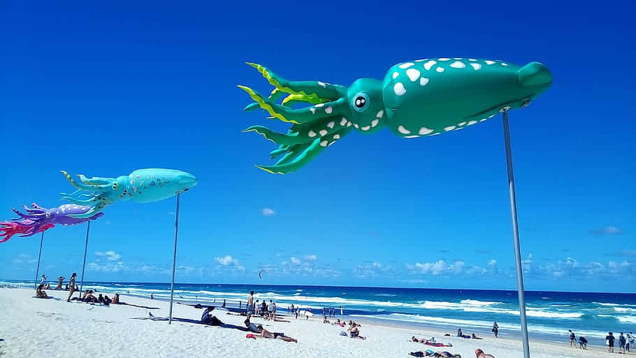  壁紙 — 色彩繽紛的巨大充氣魷魚雕塑聳立在陽光明媚的海灘上，人們正在放鬆，海浪拍打著。