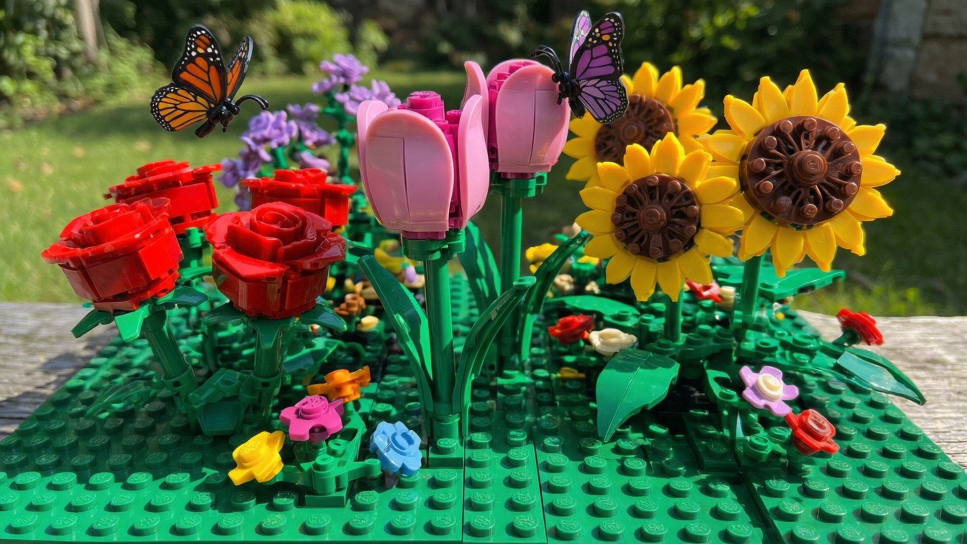 Lego Flower Garden 壁紙 — 一個充滿活力的LEGO花園，有紅玫瑰、粉紅鬱金香、黃色向日葵和蝴蝶在綠色底板上。