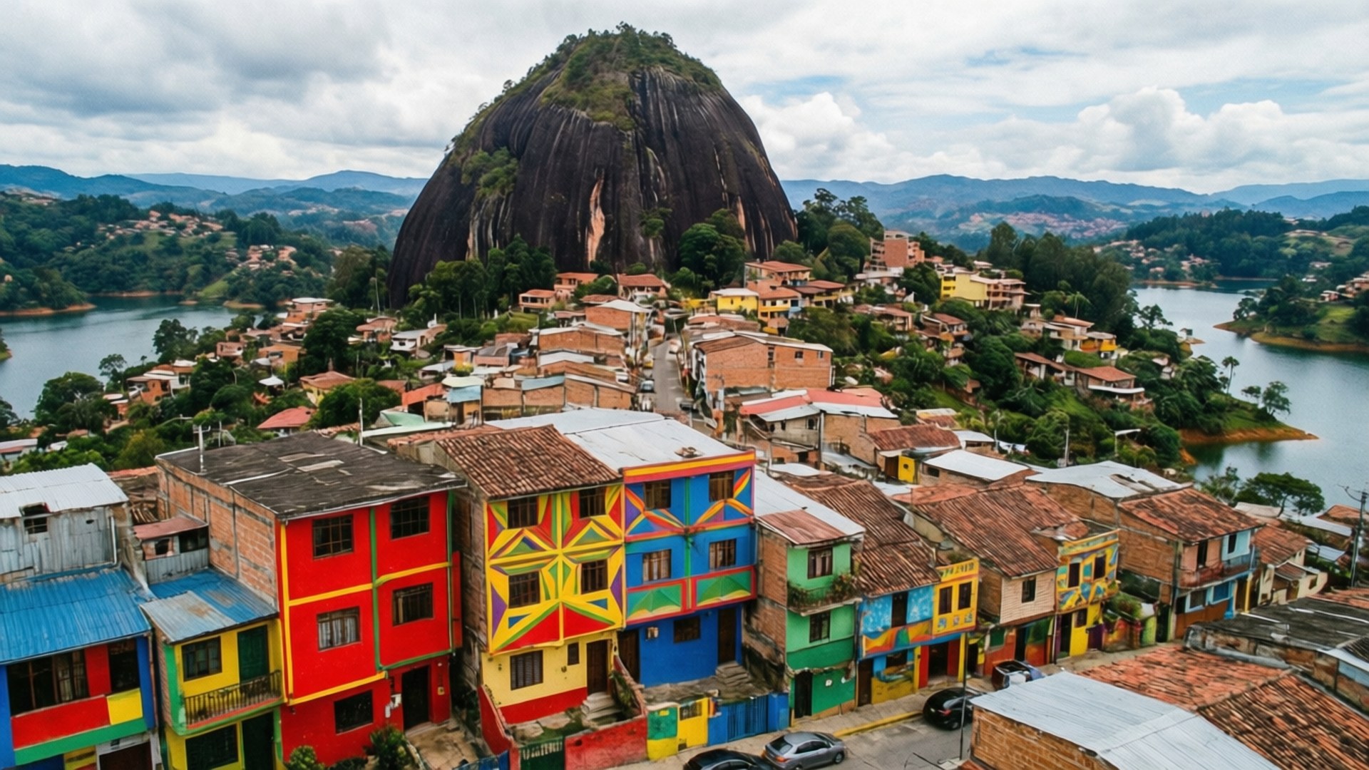 Guatapé Aerial 壁紙 — 色彩繽紛的房屋依附在巨大的深色岩石山斜坡上，俯瞰著陰天天空下平靜的湖泊。
