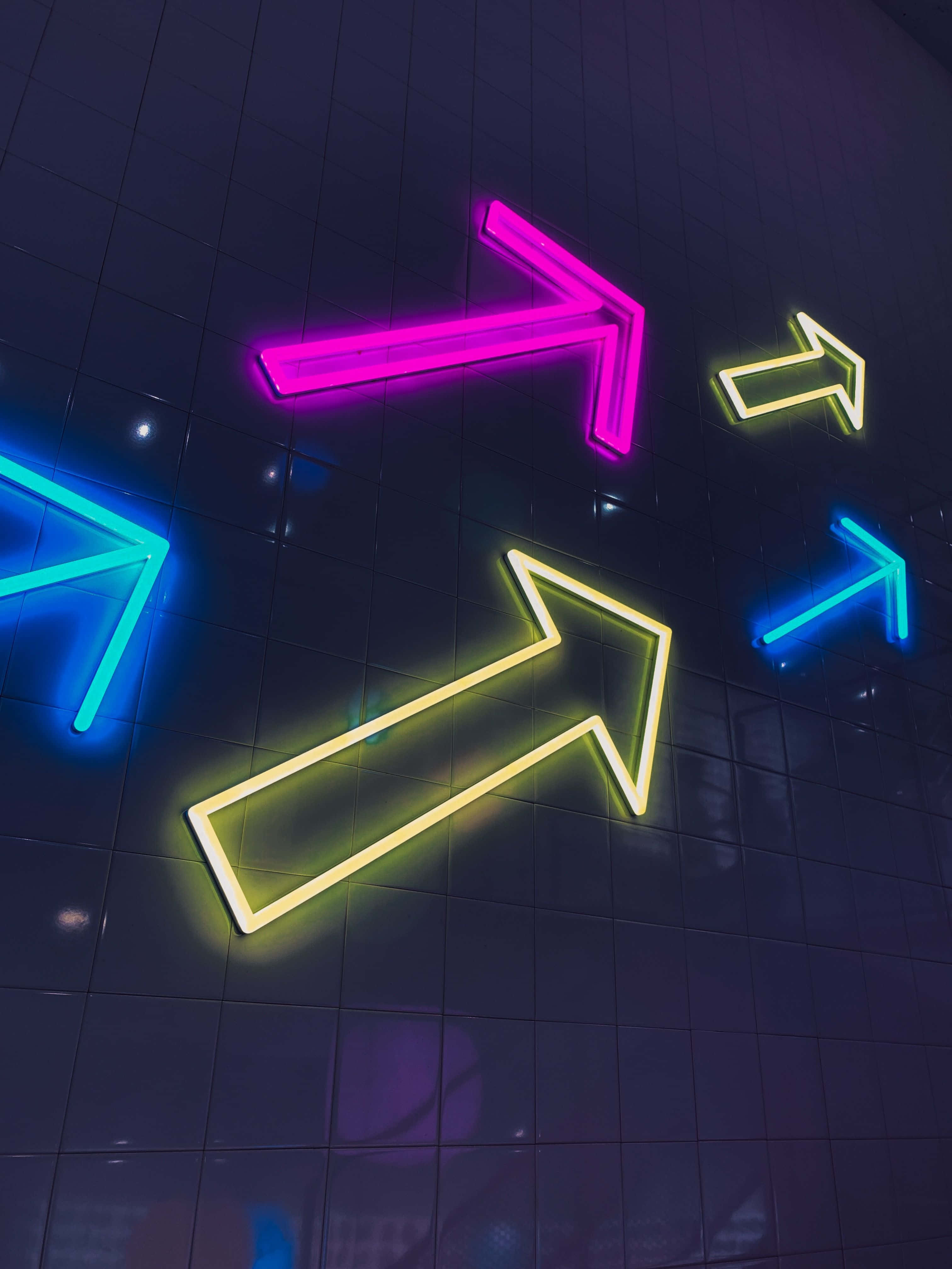 Neon Light 桌布 壁紙 — 充滿活力的粉紅色、黃色和藍色霓虹箭頭在深色瓷磚牆上發光，營造出動感未來主義的城市氛圍。