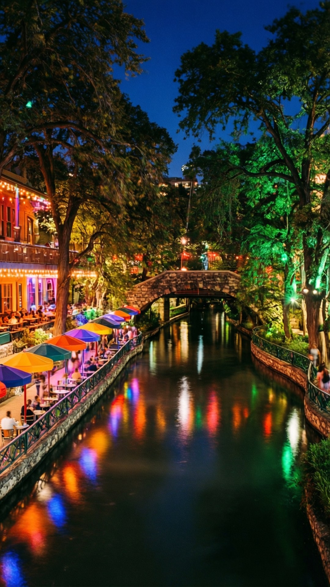 River Walk Night Lights 壁紙 — 石橋下運河旁的熱鬧夜景，色彩繽紛的雨傘在燈串下閃耀，倒影在水面上舞動。