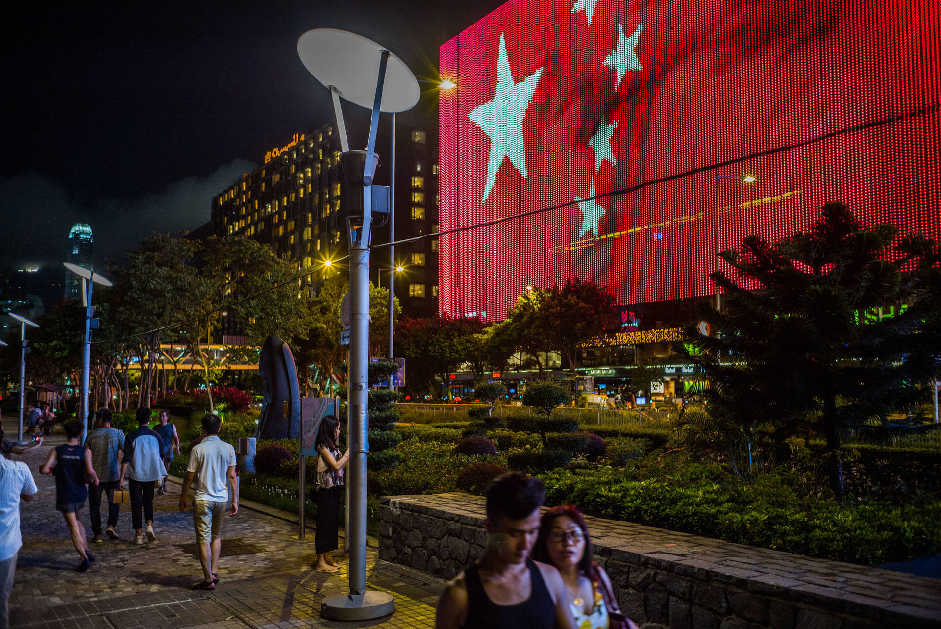 夜間城市場景，人們走過建築物上大型的旗幟燈光展示。 — 中國國旗 壁紙