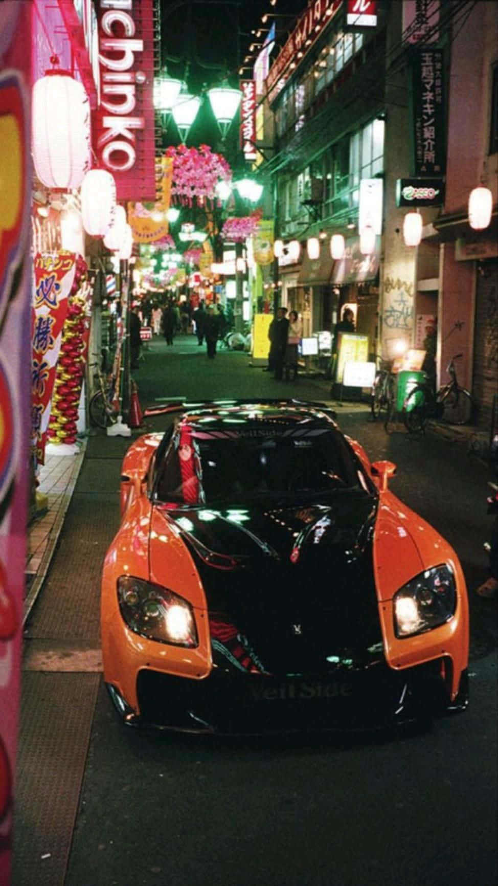  壁紙 — 一輛鮮豔的橙色和黑色跑車停泊在夜晚霓虹燈閃爍的東京小巷中，周圍有發光的招牌和燈籠。
