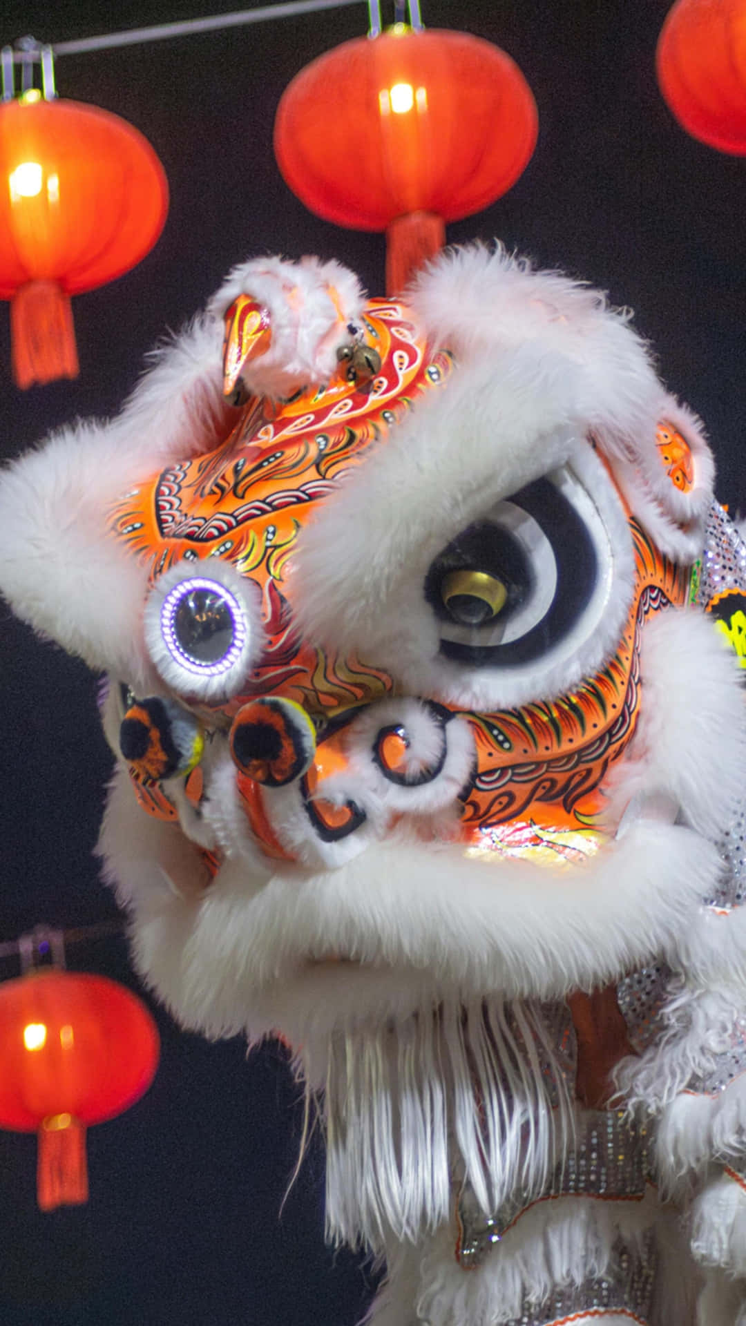  壁紙 — 一個充滿活力的橙色和白色舞獅服裝，帶有發光的眼睛和蓬鬆的白色毛髮，在黑暗背景下被紅色燈籠照亮。
