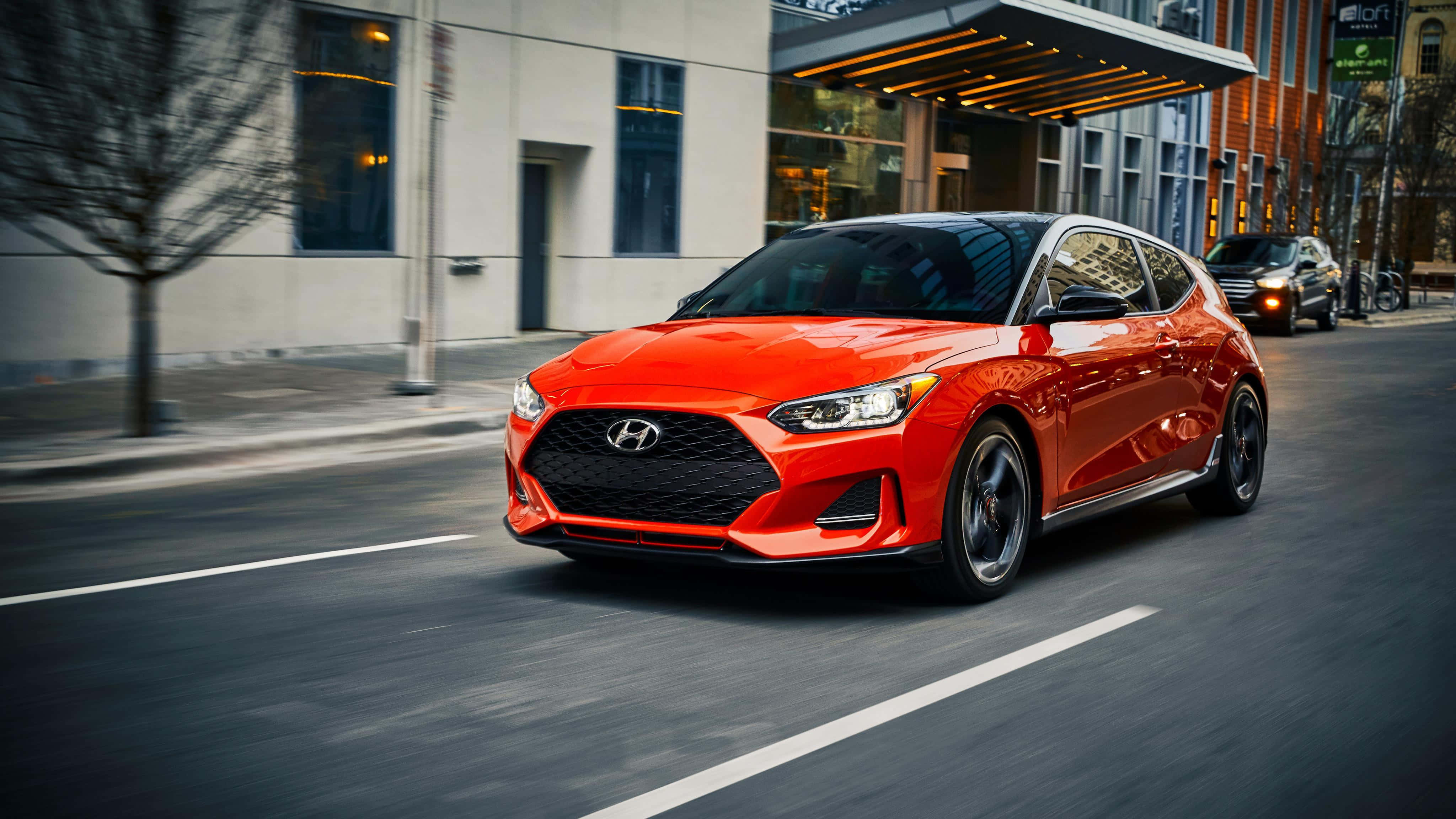 Hyundai Veloster 桌布 壁紙 — 一輛充滿活力的橙色現代Veloster在街道上疾馳，帶有動態模糊效果，背景是現代建築。