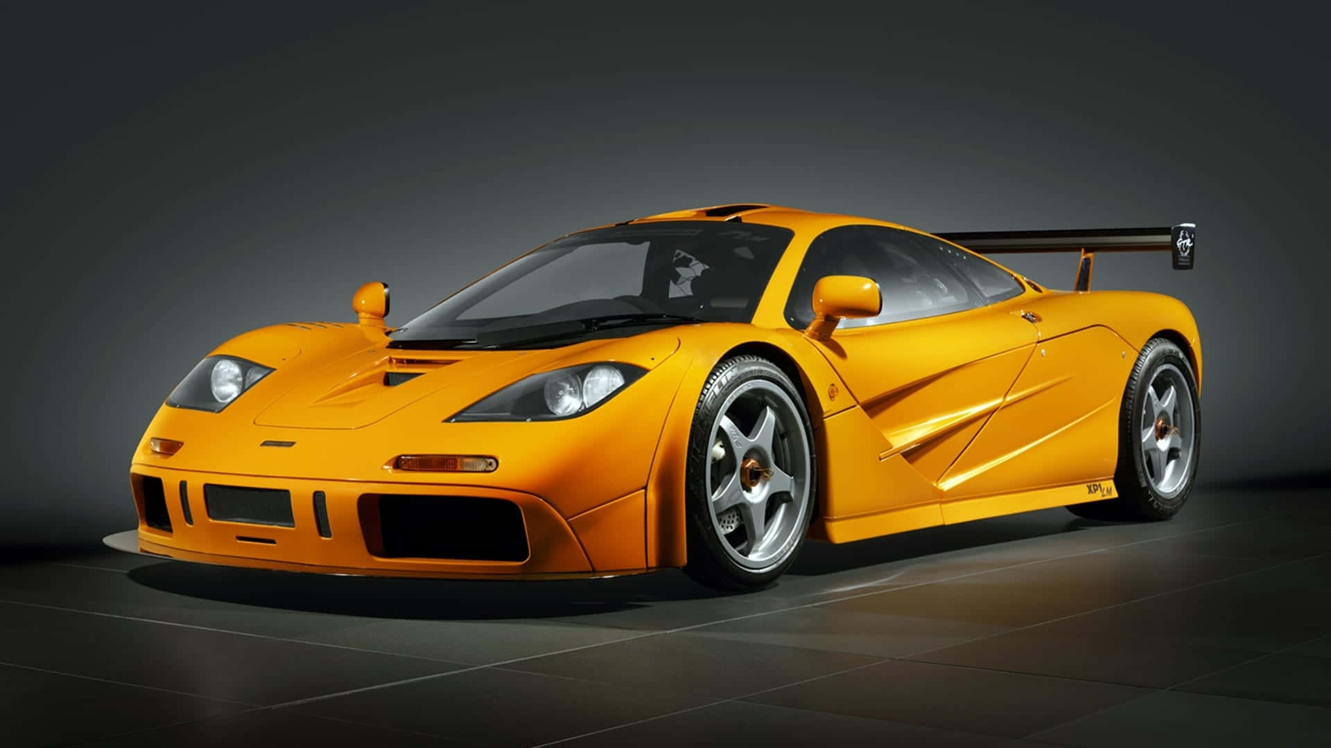 Mclaren F1 桌布 壁紙 — 一輛充滿活力的橙色McLaren F1超級跑車，配備大型尾翼，在昏暗的工作室中以戲劇性燈光展示。