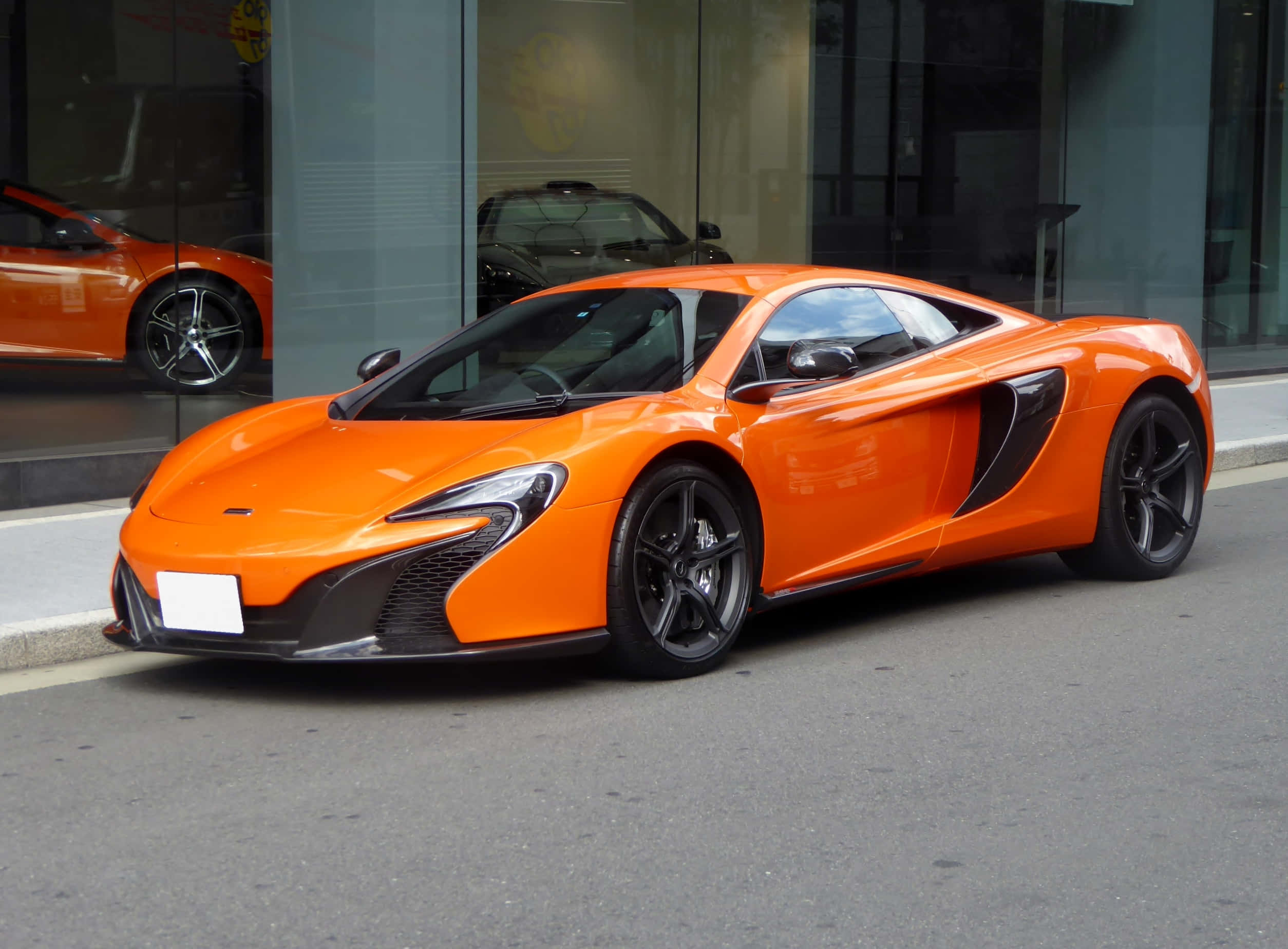 Mclaren 650s 桌布 壁紙 — 一輛充滿活力的橙色McLaren跑車在日光下閃閃發光，停放在玻璃建築旁的城市街道上。
