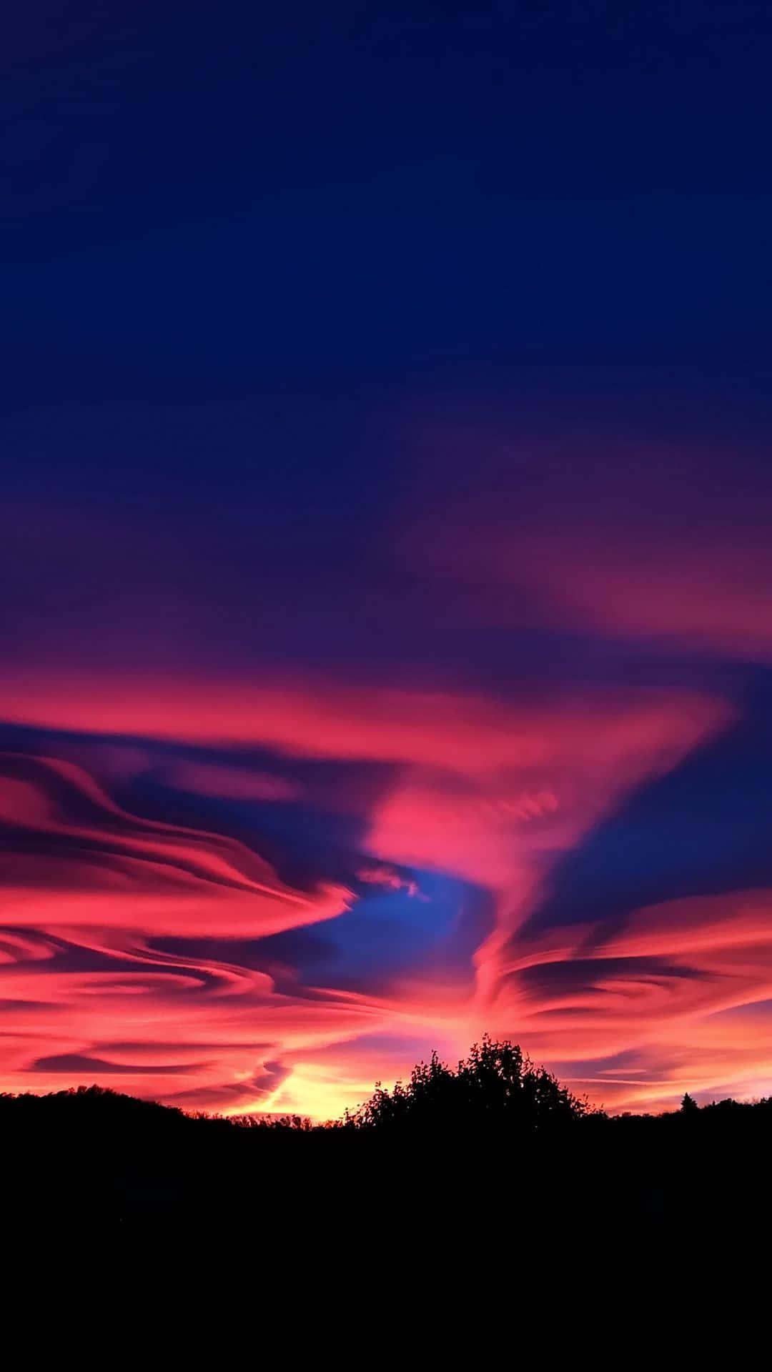 Colorful Sunset 桌布 壁紙 — 鮮豔的粉紅色和紫色雲彩在深藍色暮色天空中戲劇性地旋轉，上方是黑暗的樹線。