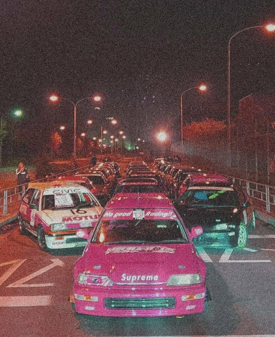 帶有 Supreme 標誌的粉紅色賽車在夜晚與其他改裝車一起停放。 — JDM汽車 壁紙