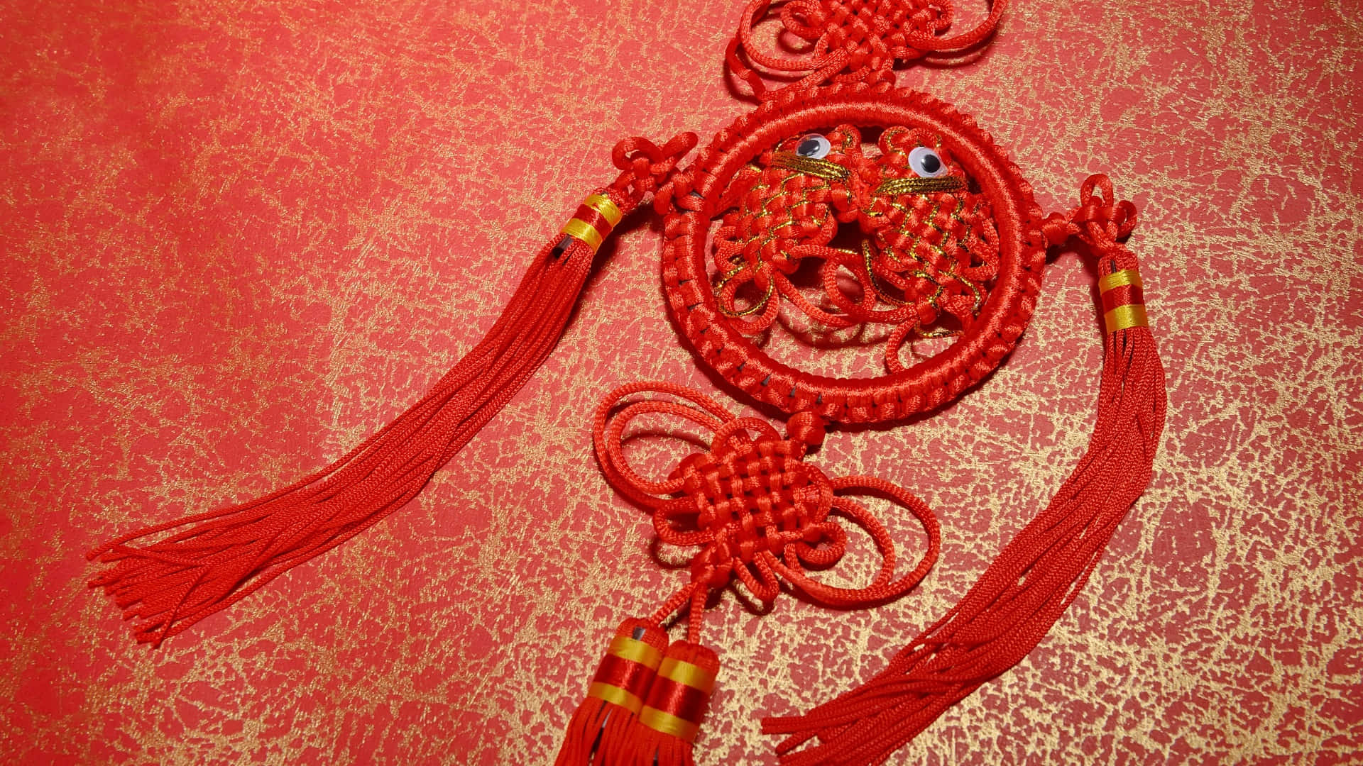  壁紙 — 一個帶有咕嚕眼和流蘇的鮮紅色中國結飾品，放置在帶有紋理的紅色和金色布料背景上。