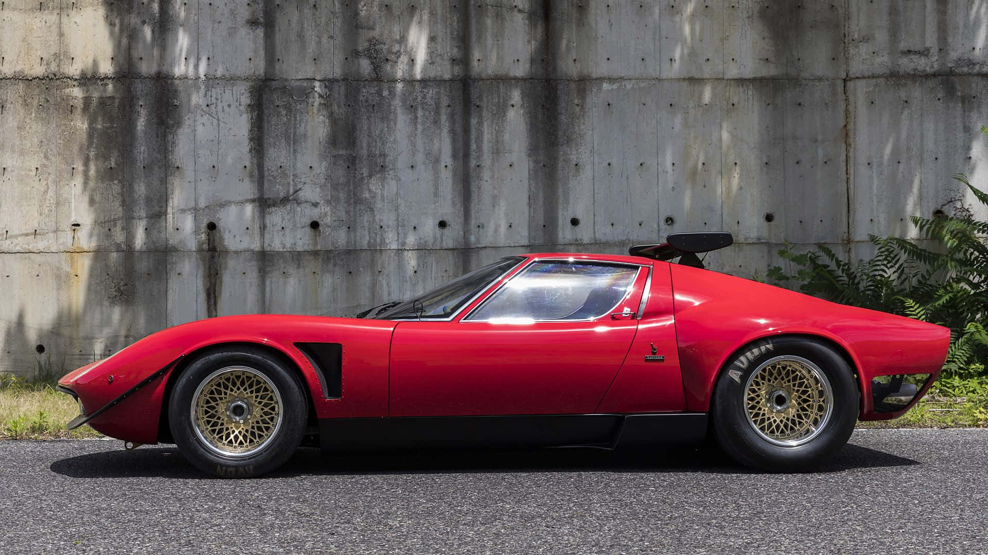 Lamborghini Miura 桌布 壁紙 — 一輛鮮紅色經典跑車配金色鋼絲輪圈，停靠在風化的混凝土牆邊。