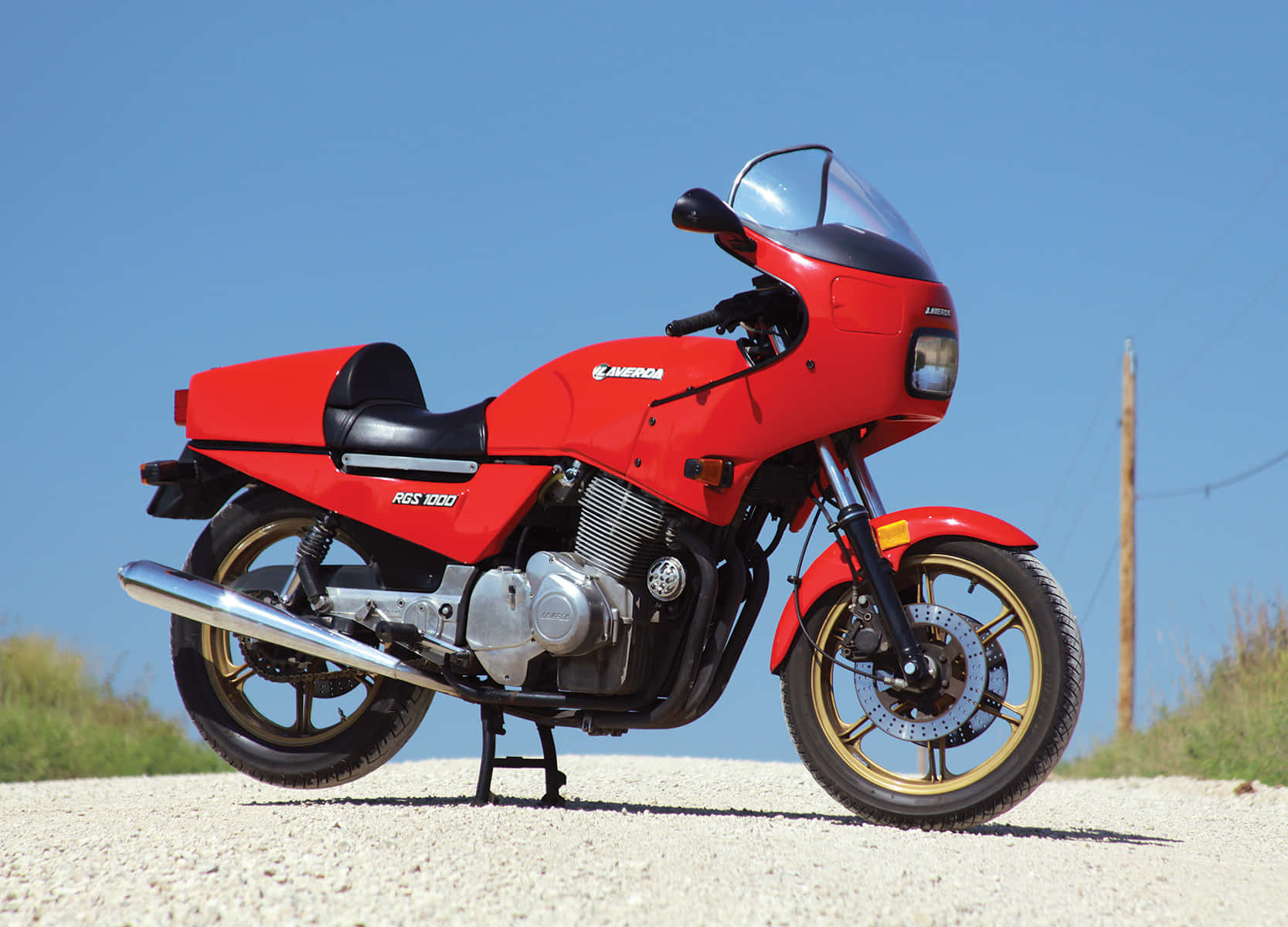  壁紙 — 一輛鮮紅色的Honda RGS 1000摩托車在晴朗藍天下的碎石路上蓄勢待發，背景有綠色草地。