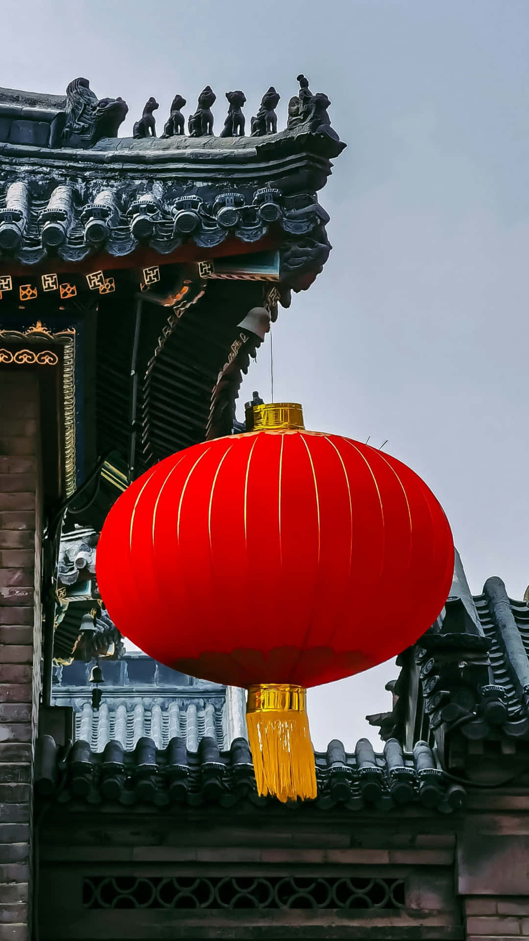  壁紙 — 一個鮮紅色的燈籠帶有金色流蘇，在陰天背景下懸掛在傳統中式瓦屋頂前，格外顯眼。