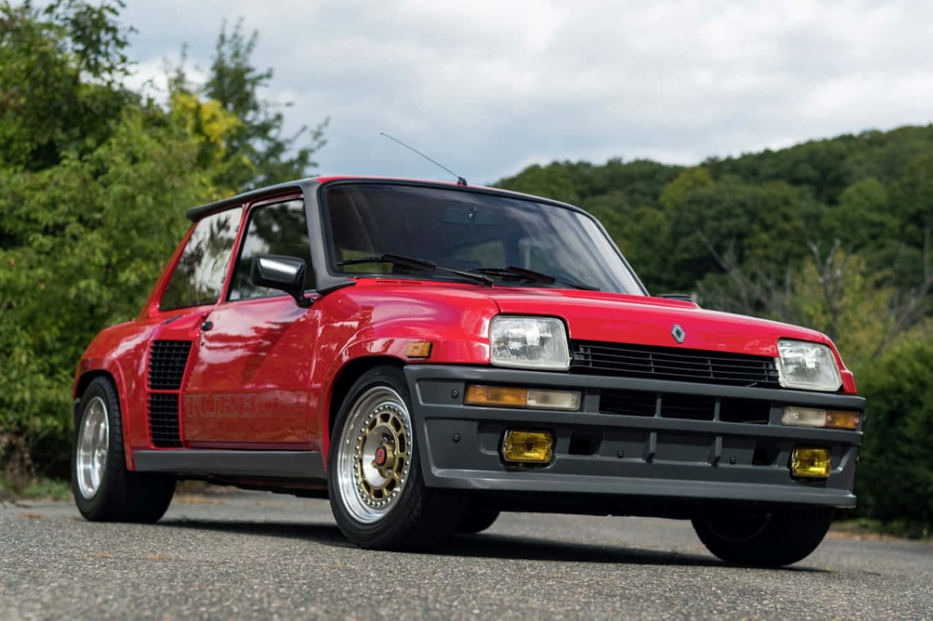 Renault 5 Turbo 桌布 壁紙 — 一輛鮮紅色的雷諾5 Turbo 2跑車停泊在碎石路上，背景是茂密的綠樹和山丘，天空多雲。