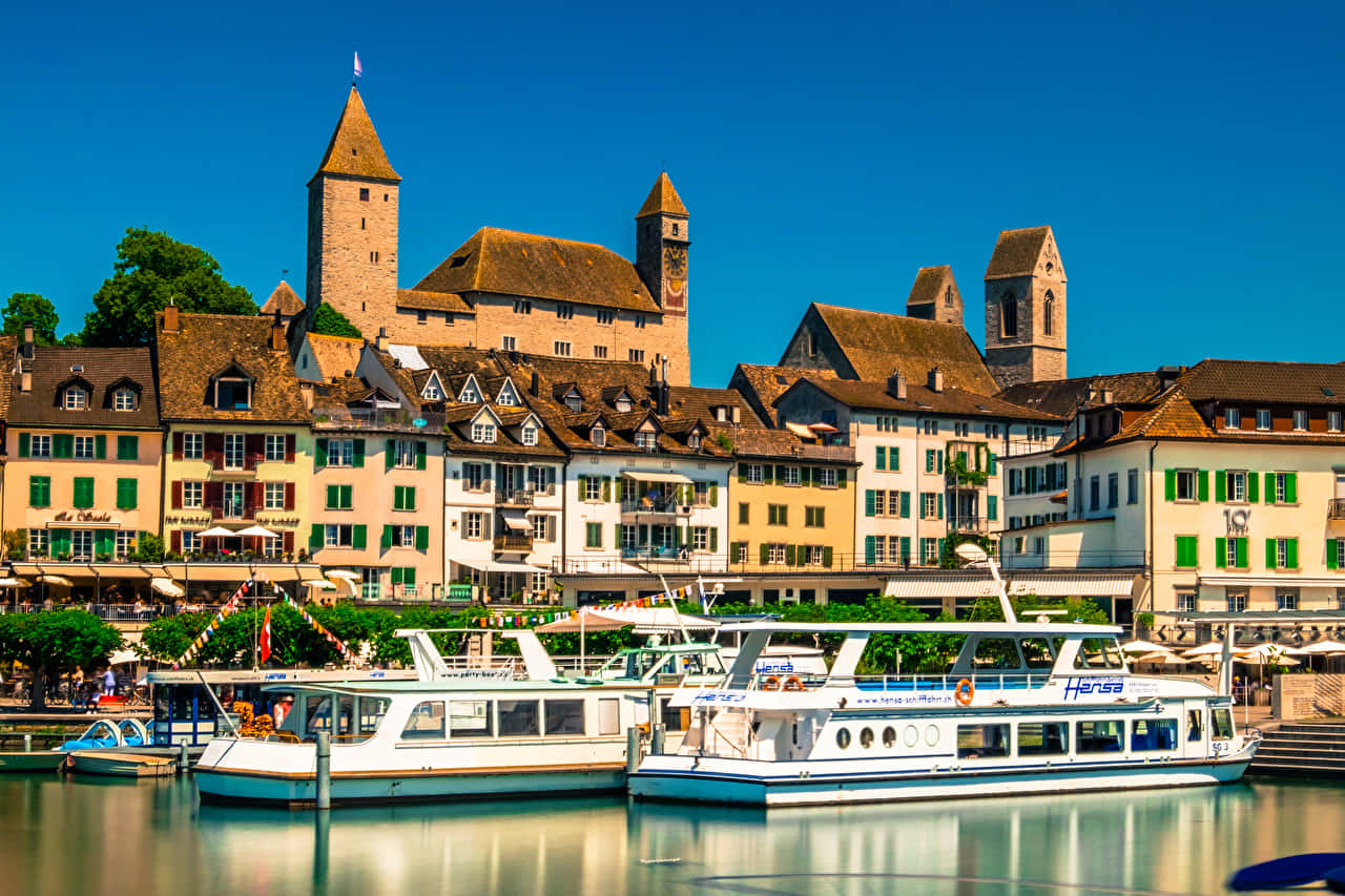 Rapperswil Jona 桌布 壁紙 — 一個充滿活力的河畔小鎮，擁有歷史悠久的石塔和色彩繽紛的建築，在晴朗的藍天下倒映在平靜的水面上。