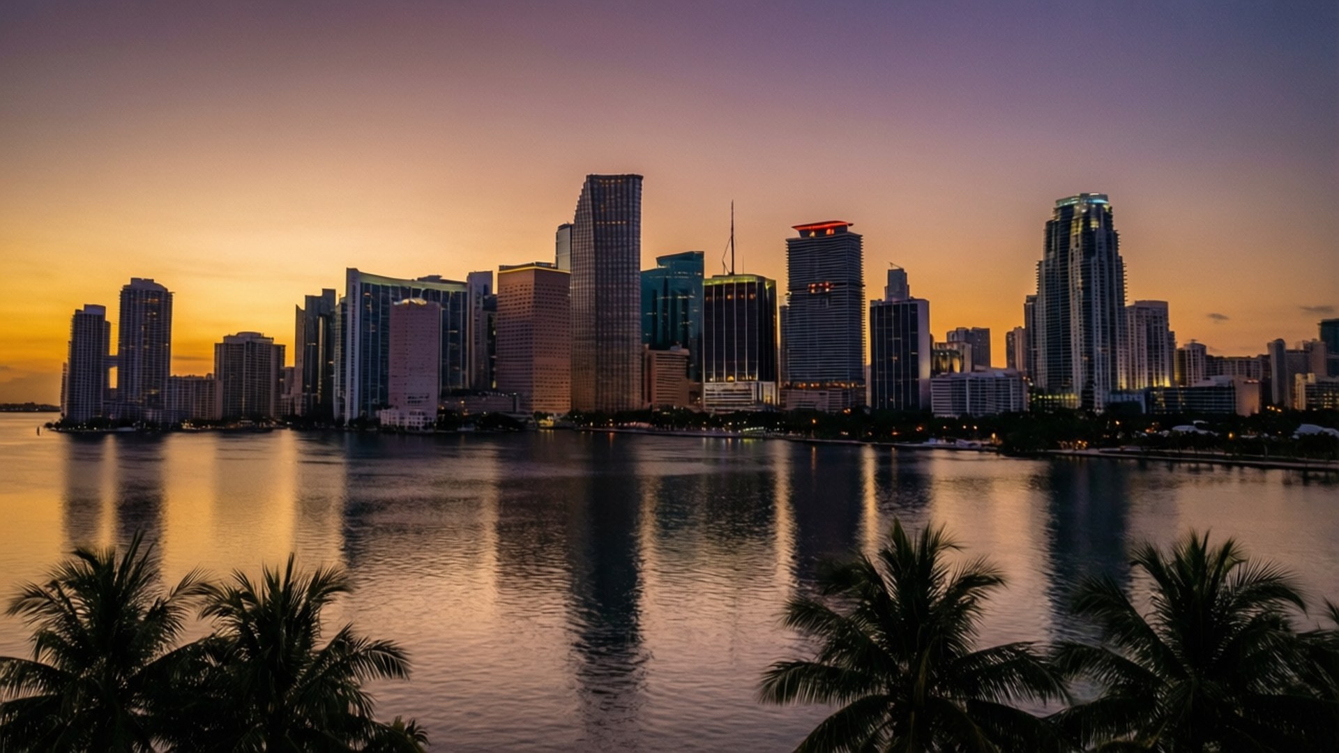 Bayfront Twilight Skyline 壁紙 — 充滿活力的夕陽將溫暖的色調投射在現代城市天際線上，倒映在平靜的水面上，並以棕櫚樹的剪影為框。