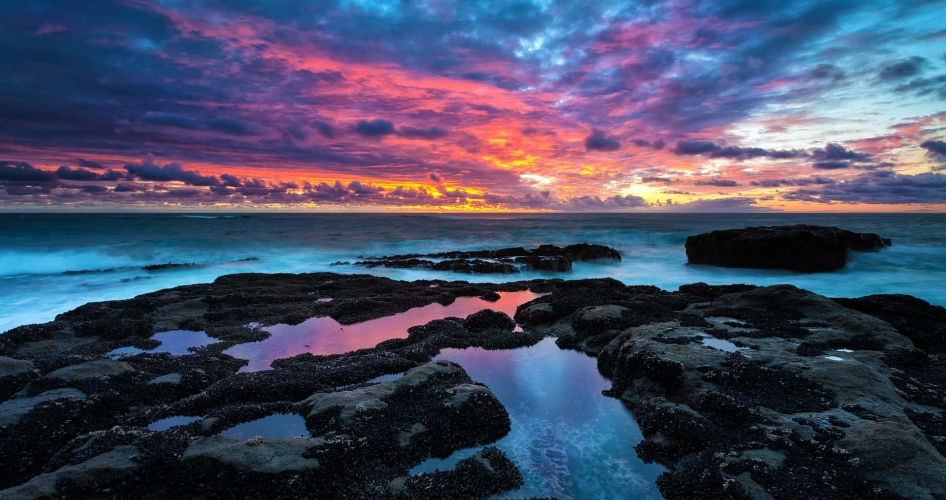 Colorful Sunset 桌布 壁紙 — 充滿活力的日落用火熱的粉紅色和紫色描繪天空，在岩石海岸線上方，有碧綠的海水和反射池。