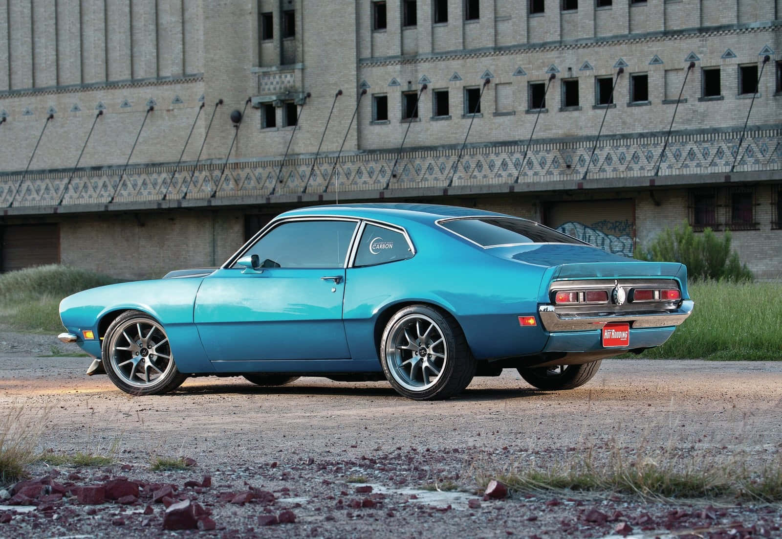 Ford Maverick 桌布 壁紙 — 一輛充滿活力的綠松石色經典肌肉車停在碎石上，前面是一座帶有塗鴉的風化工業建築。