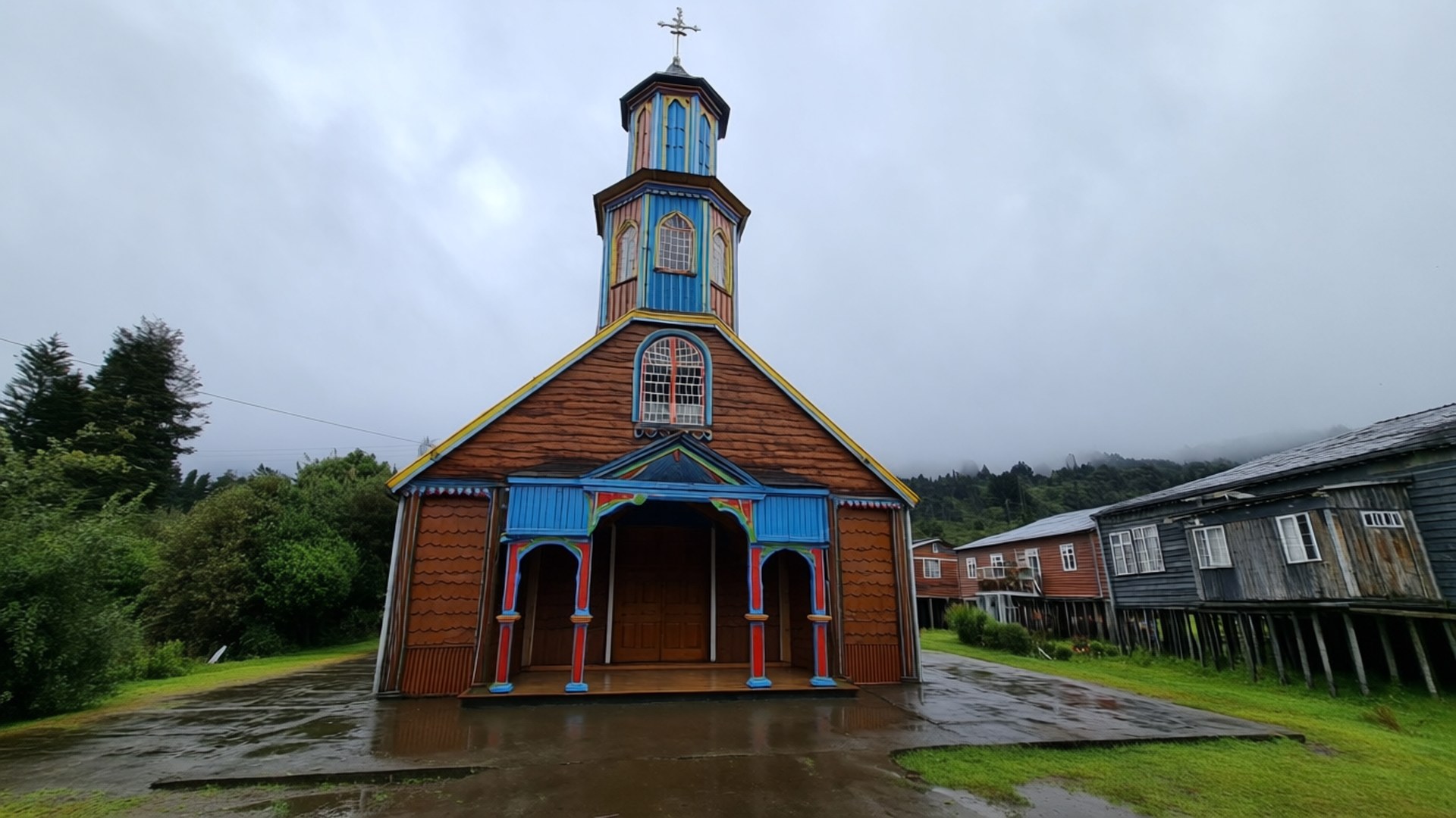Chiloé Wooden Church 壁紙 — 一座充滿活力的木造教堂，帶有藍色和紅色點綴，矗立在灰色的天空下，周圍是潮濕的草地和質樸的木屋。
