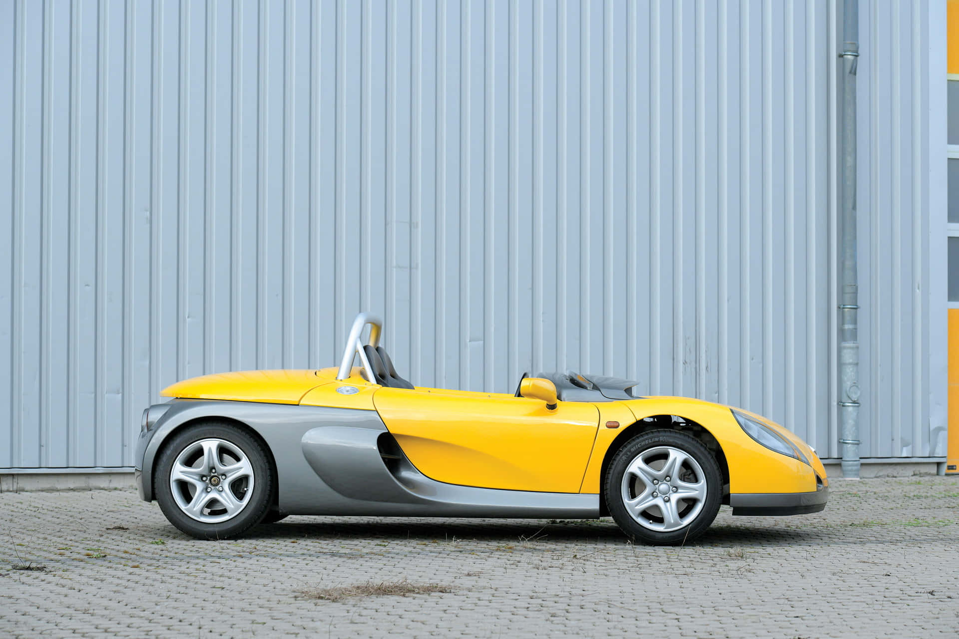 Renault Sport Spider 桌布 壁紙 — 一輛鮮黃色和銀色的跑車停放在鋪砌路面上，靠著波紋金屬牆。