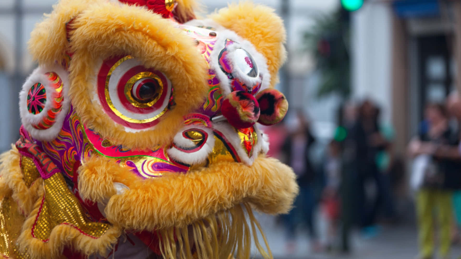  壁紙 — 鮮黃色舞獅服裝，帶有複雜的紅色、金色和粉紅色圖案，在模糊的城市街景中脫穎而出。