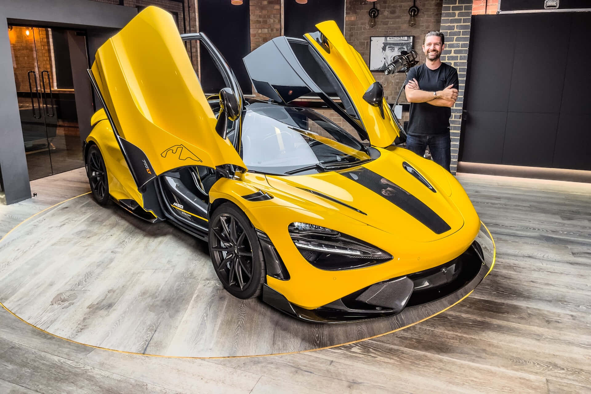 Mclaren 765lt 桌布 壁紙 — 一輛鮮黃色McLaren跑車打開蝴蝶門，停在圓形木質平台上，一位微笑的男子在現代車庫中站在旁邊擺姿勢。