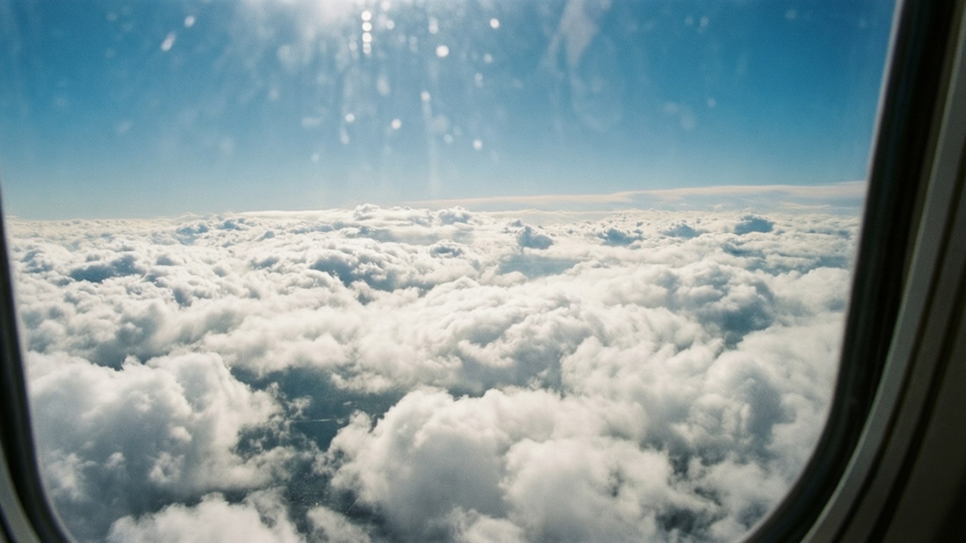 Above the Clouds 壁紙 — 從飛機窗戶看到的景色，無盡的蓬鬆白雲在明亮的藍天下，帶有太陽耀斑。
