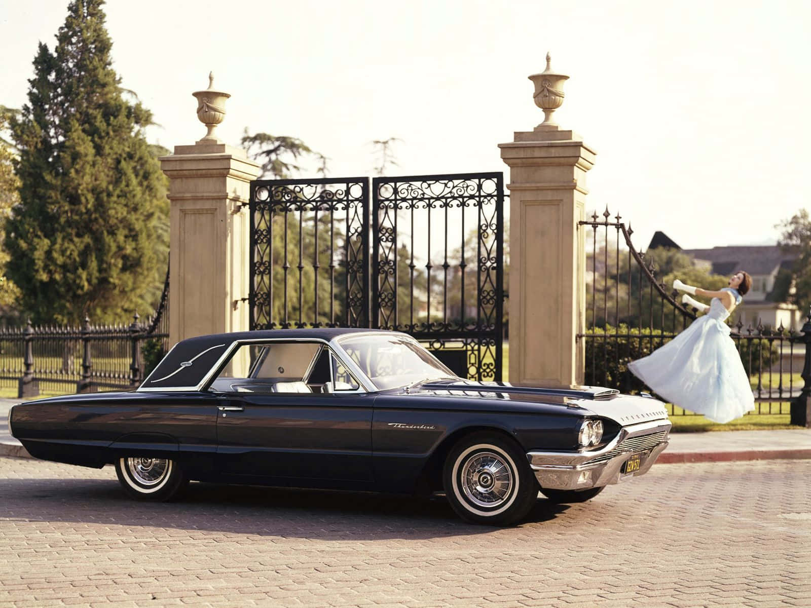 Ford Thunderbird 桌布 壁紙 — 一輛復古黑色雷鳥在華麗的大門旁閃閃發光，一位身穿飄逸白色連衣裙的女子在陽光普照的車道上歡快地跳躍。