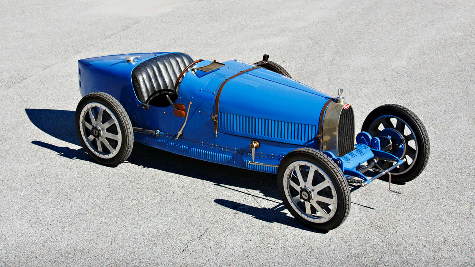 Bugatti Type 41 Royale 桌布 壁紙 — 一輛復古藍色Bugatti賽車，配備黑色皮革座椅和銀色輪圈，停放在陽光照射的混凝土表面上。
