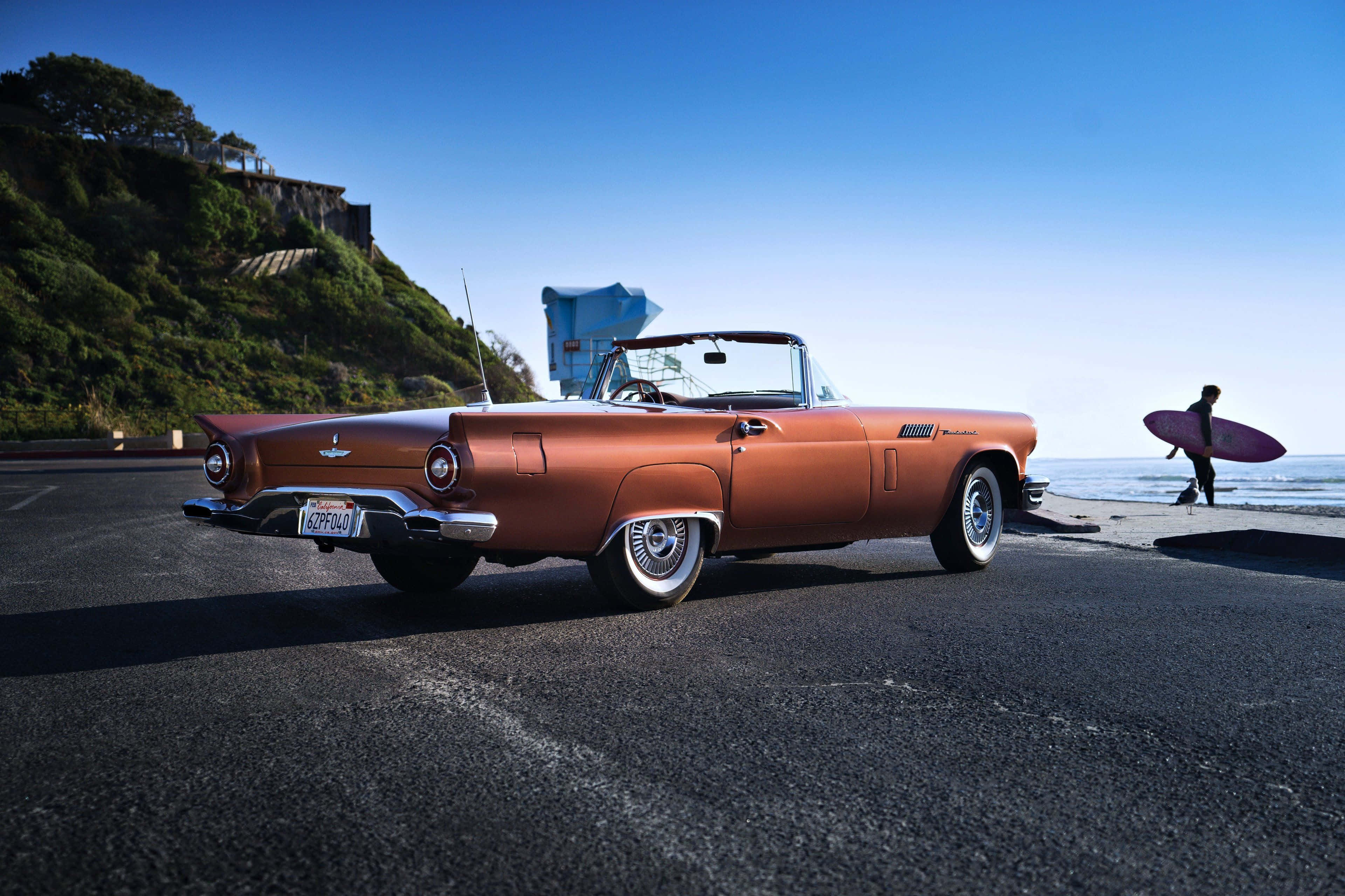 Ford Thunderbird 桌布 壁紙 — 一輛復古銅色敞篷車停泊在海岸公路上，一名衝浪者在晴朗的藍天下走向海洋。