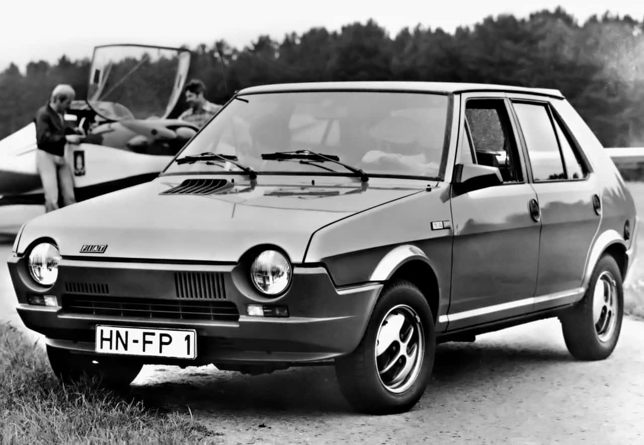 Fiat Ritmo 桌布 壁紙 — 一輛復古飛雅特126p汽車停泊在泥路上，背景有兩名男子靠近一架小型飛機。