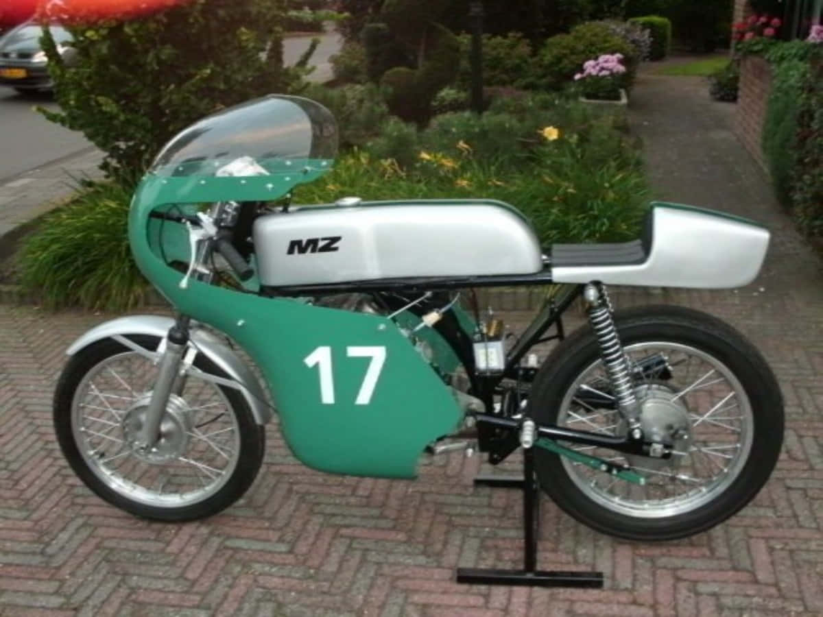 Mz Motorcycle 桌布 壁紙 — 一台復古綠色和銀色MZ賽車摩托車，號碼17，停放在磚砌車道上，周圍環繞著茂密的花園葉子。